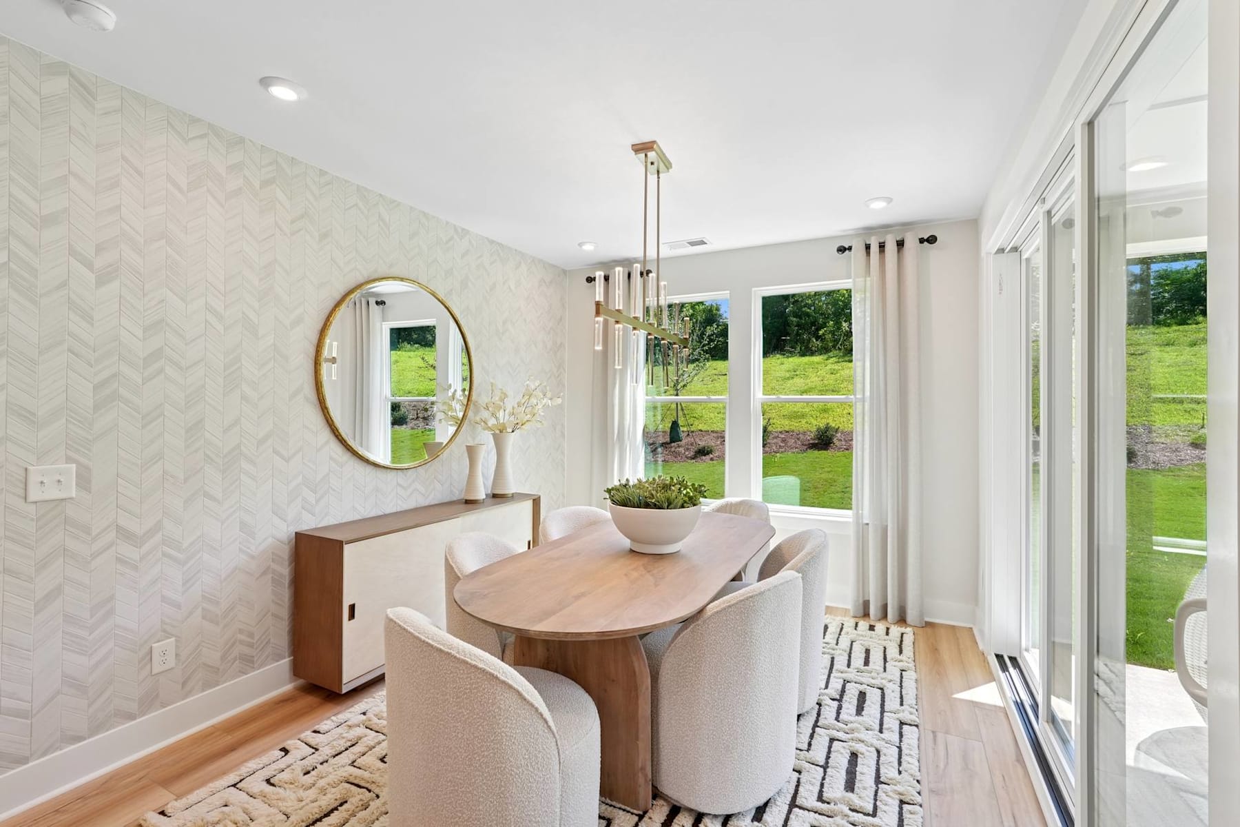 A modern and minimalist dining area with a wooden table, chairs, and a decorative mirror on the wall, overlooking a scenic outdoor landscape through large windows.