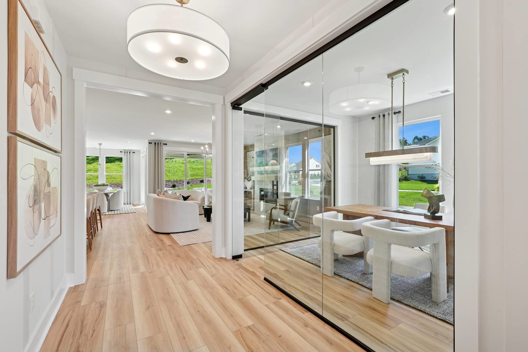 A bright and spacious interior with a wooden floor, white walls, and large windows overlooking a scenic outdoor landscape. The room features modern furniture and decor, including a round light fixture and a mirrored wall that creates a sense of depth and openness.