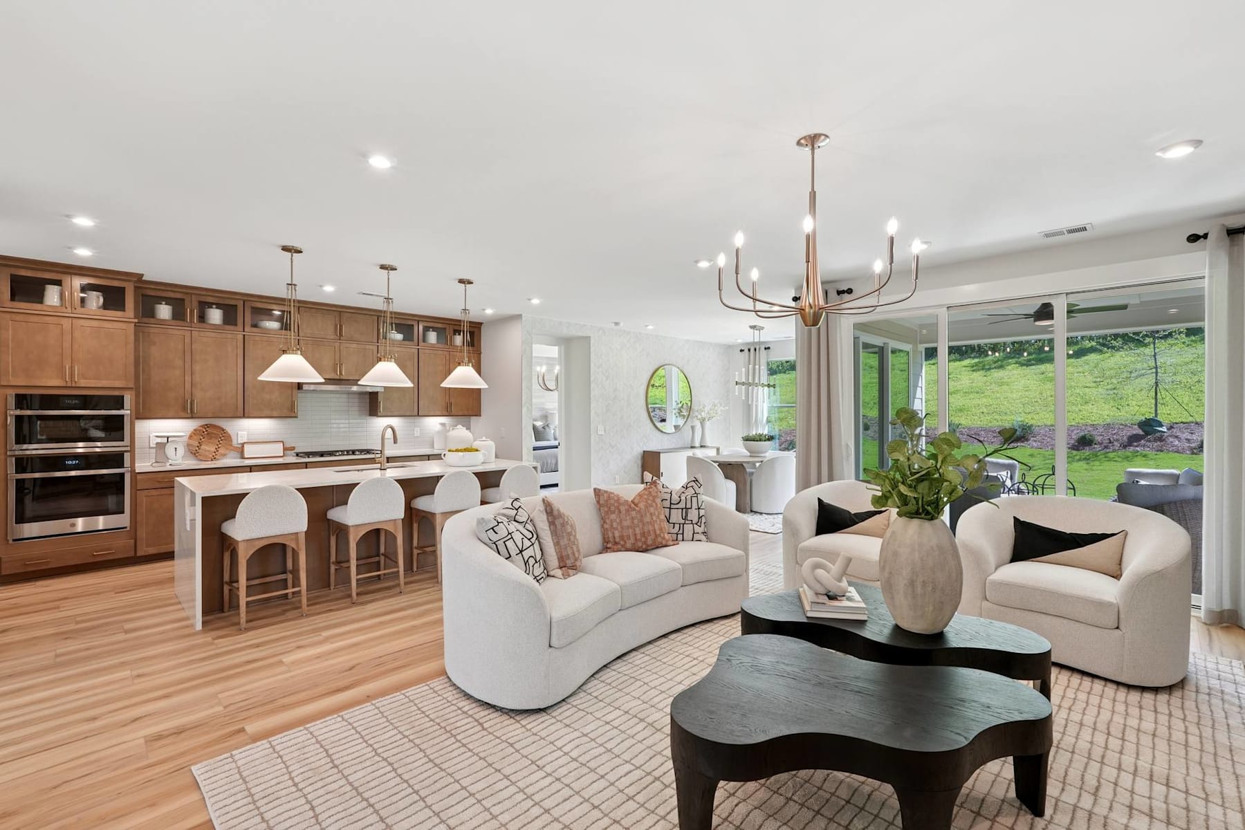 A spacious and modern open-concept living room with a kitchen, dining area, and comfortable seating arrangements, surrounded by large windows overlooking a lush outdoor landscape.