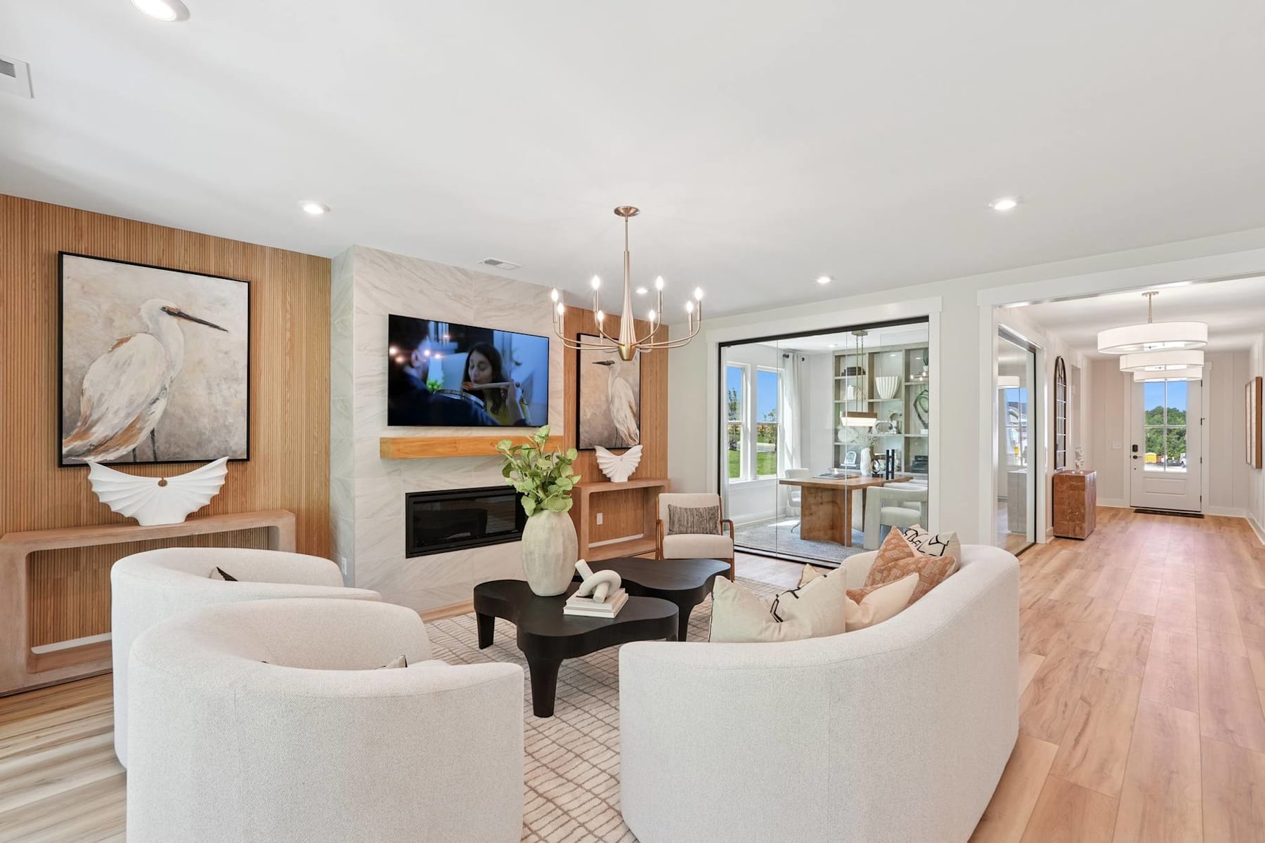 A modern and spacious living room with white furniture, a large TV, and a wooden accent wall, leading to an open doorway with a view of the outdoors.