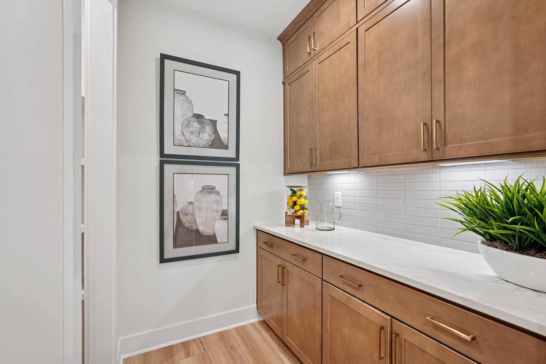 The image depicts a modern kitchen with wooden cabinets, white countertops, and framed artwork on the walls.