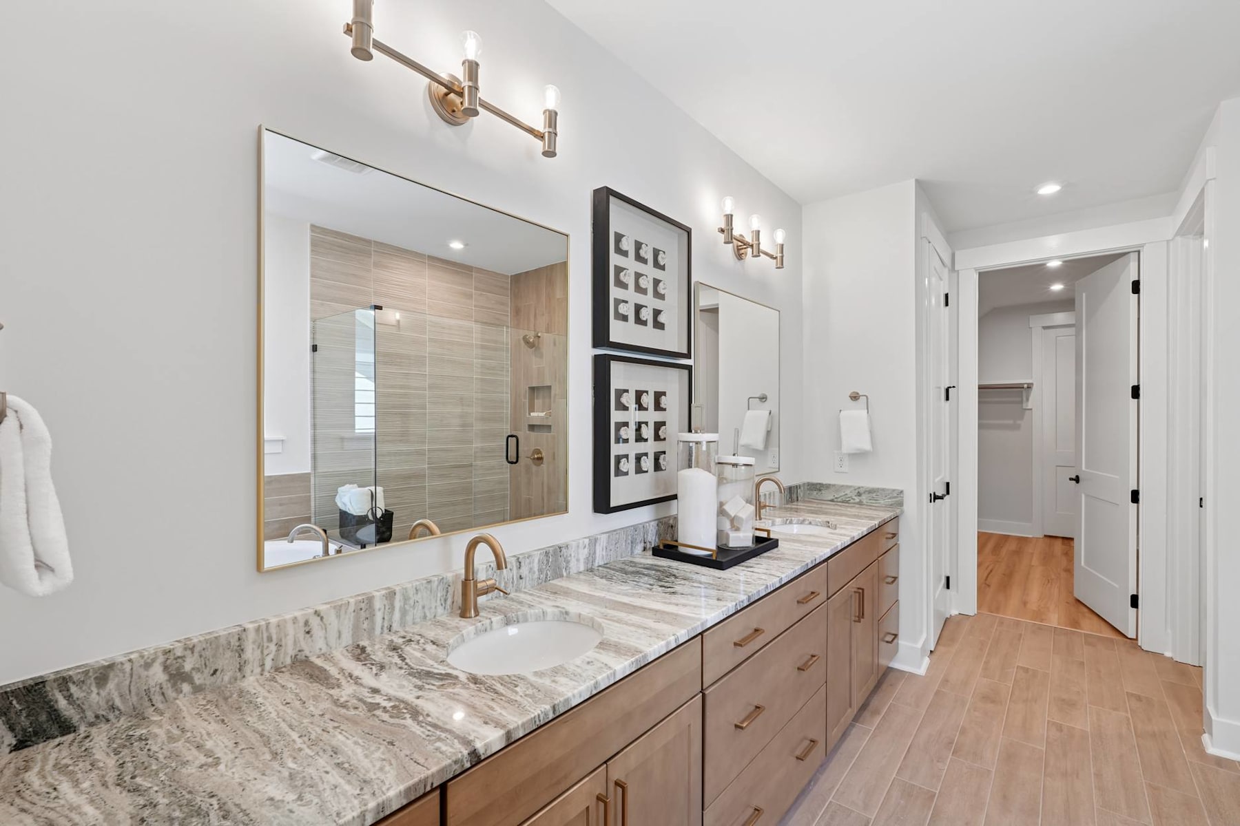 A modern and well-designed bathroom with a large vanity, a mirror, and a hallway visible in the background. The vanity features a marble countertop and wooden drawers, complemented by wall sconces and a framed artwork.