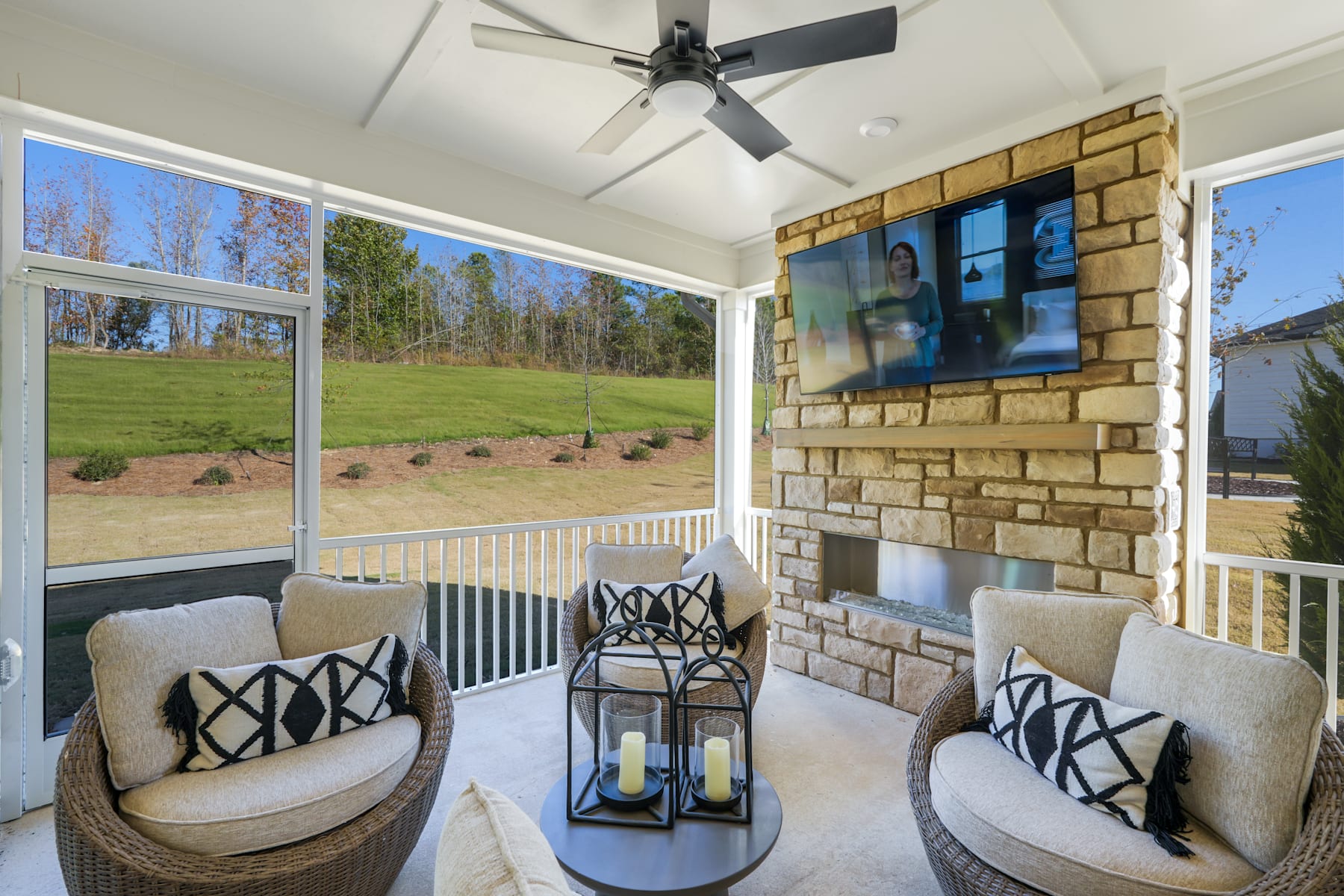 A cozy outdoor patio with comfortable seating, a stone fireplace, and large windows overlooking a grassy field and wooded area.