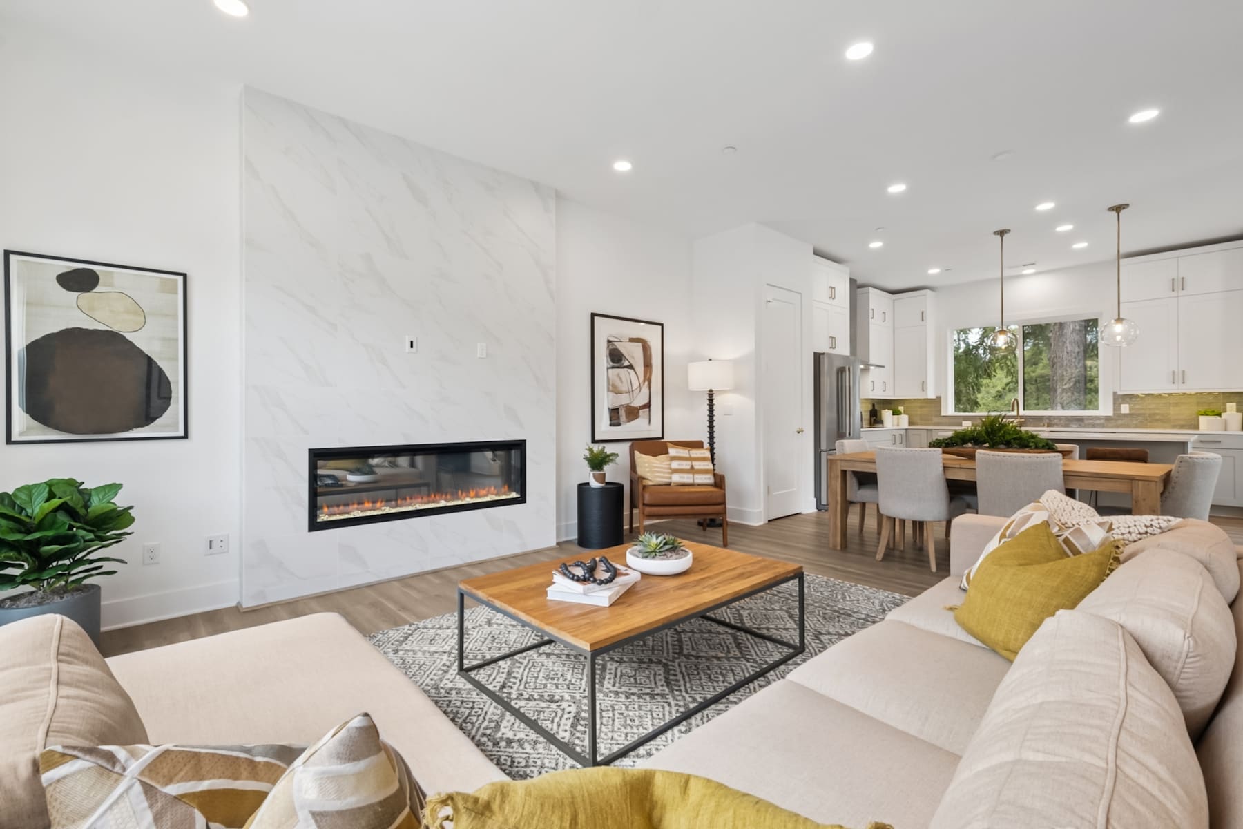 A modern and minimalist living room with a fireplace, a wooden coffee table, and a couch with yellow and white pillows, set against a backdrop of a kitchen area with white cabinets and a dining table.