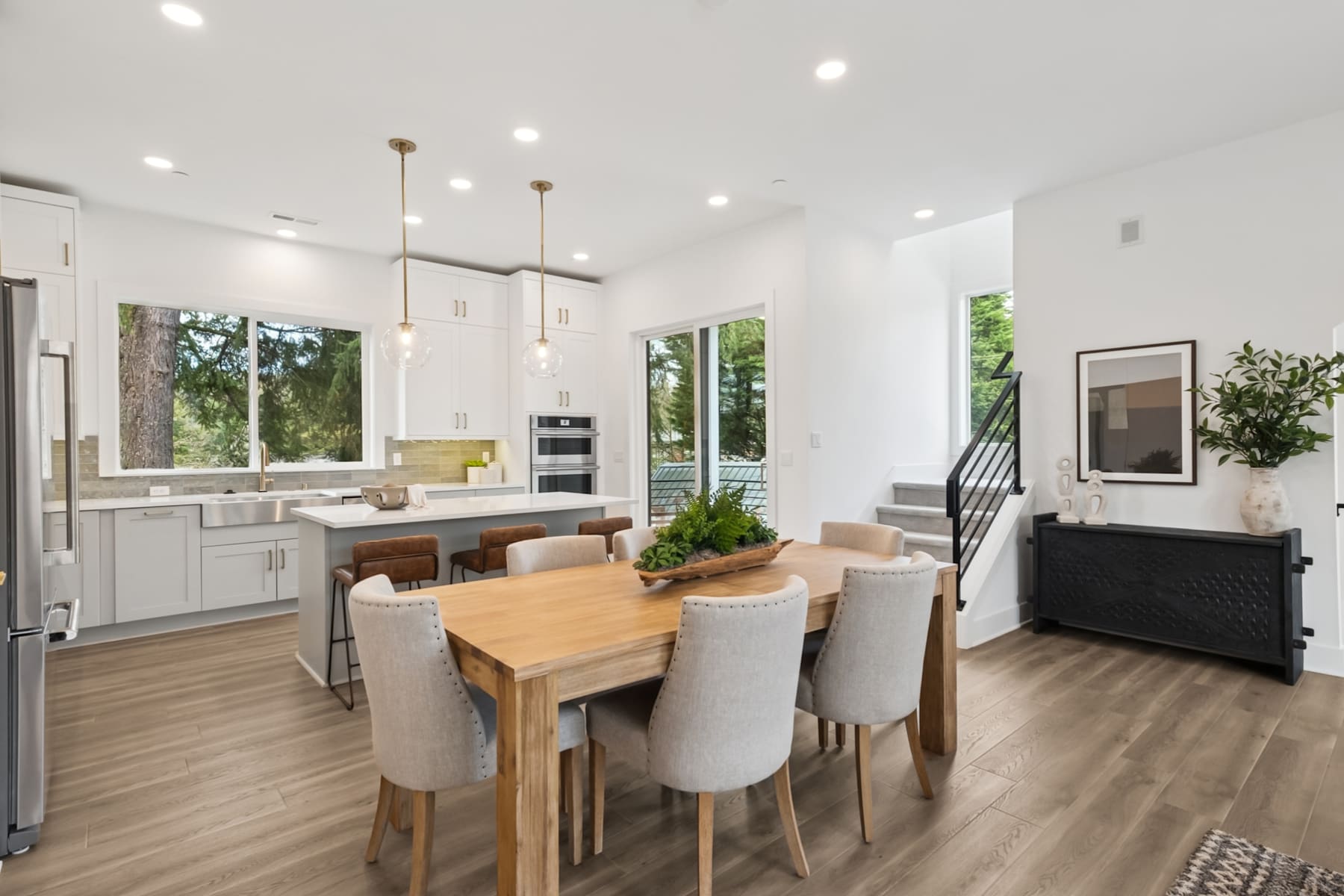 A bright and airy open-concept kitchen and dining area with white cabinets, wooden dining table and chairs, and large windows overlooking greenery outside.