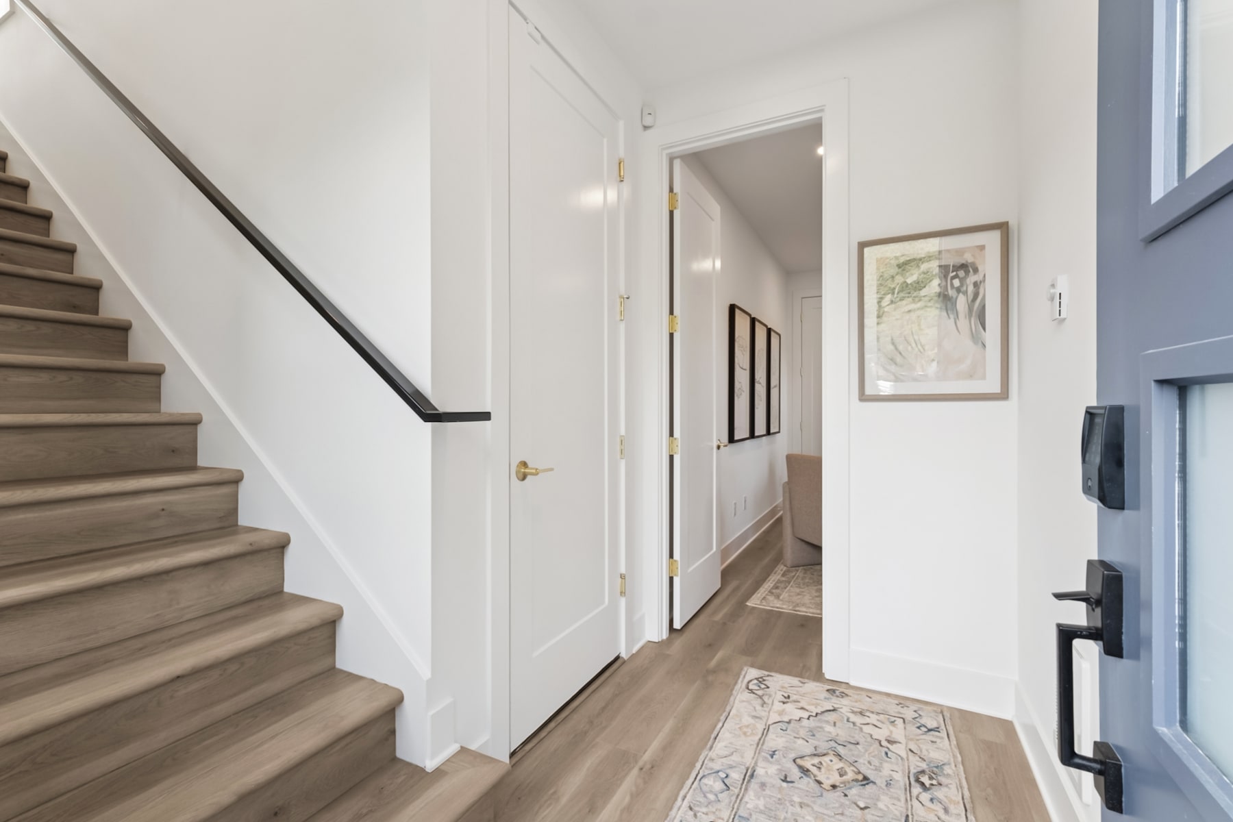 A well-lit, modern hallway with wooden stairs leading up, a framed artwork on the wall, and a doorway visible in the background.
