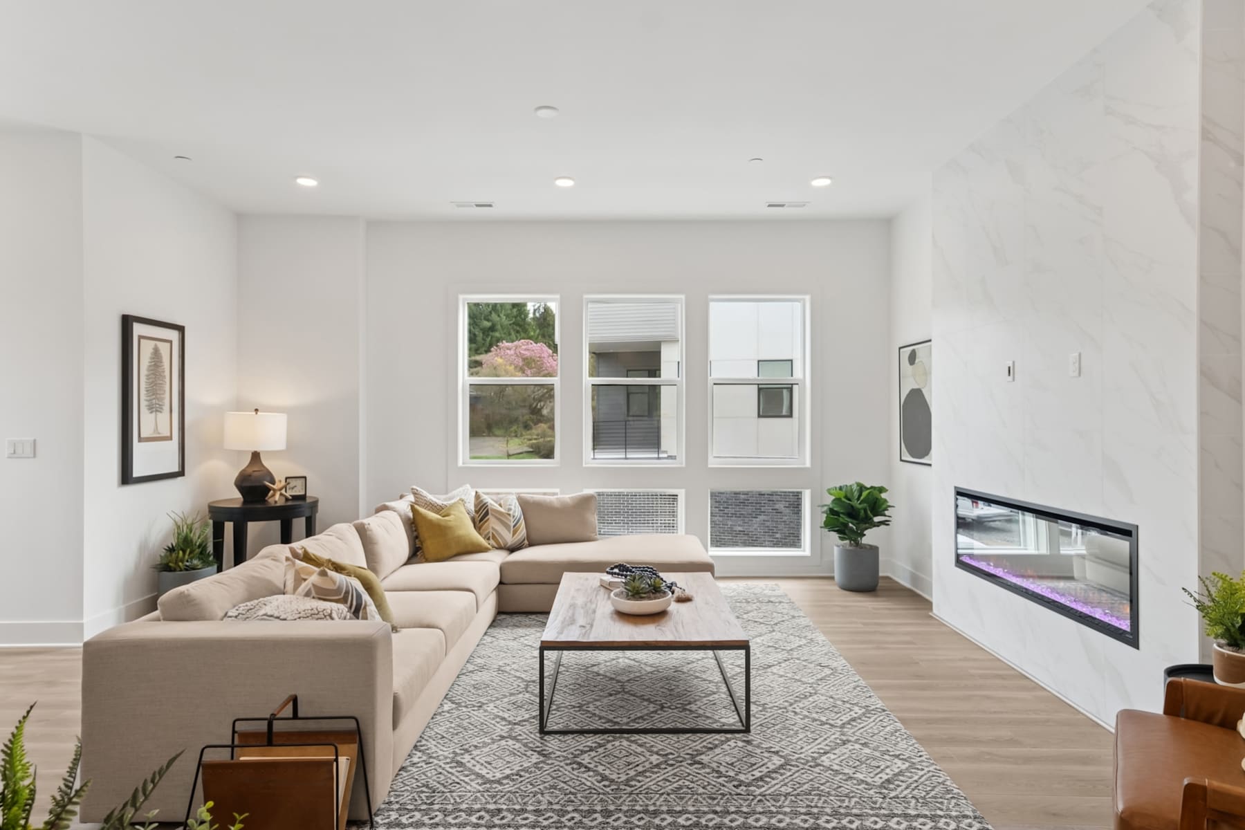 A cozy and modern living room with a comfortable beige sectional sofa, a glass-topped coffee table, and various decorative elements such as plants and artwork on the walls.