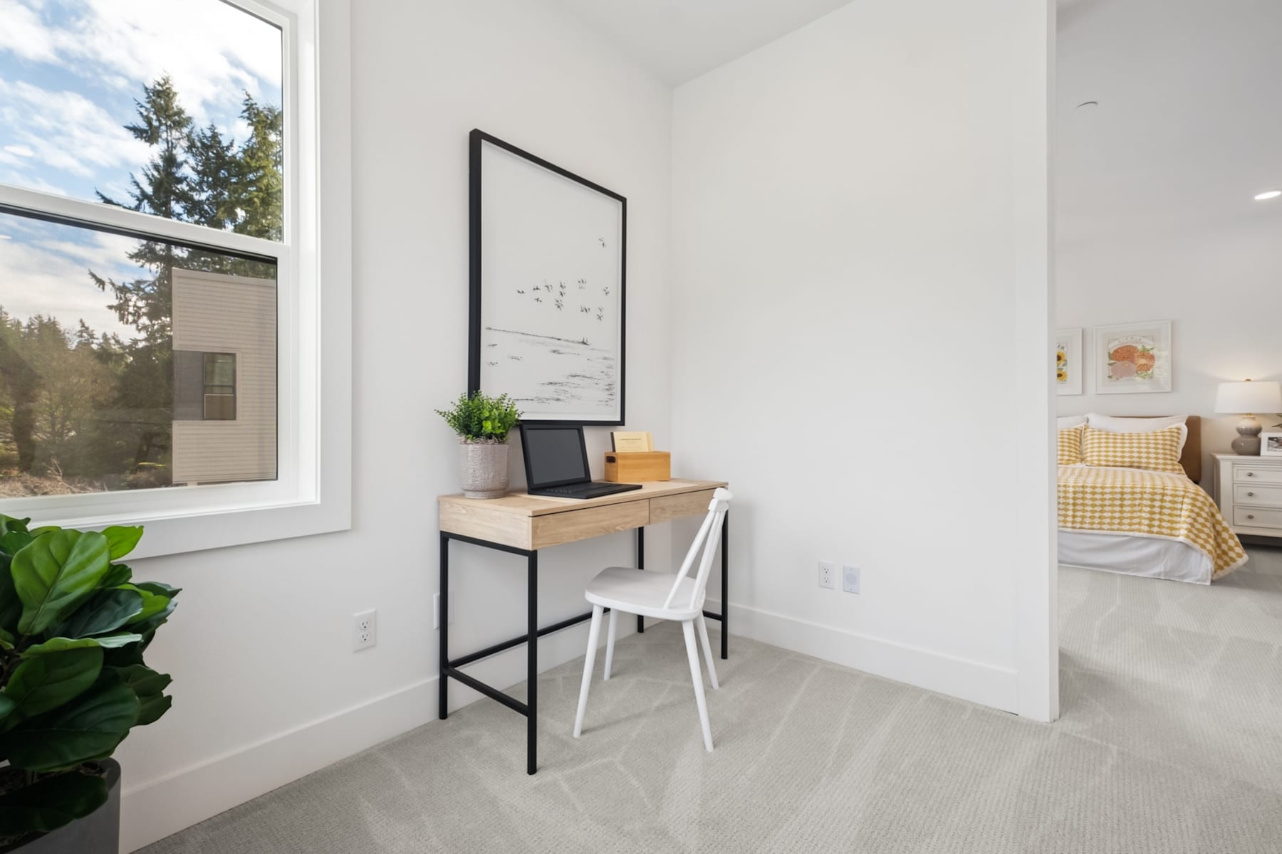A minimalist home office setup with a wooden desk, a white chair, and a framed artwork on the wall, set against a backdrop of a large window overlooking a forested area.