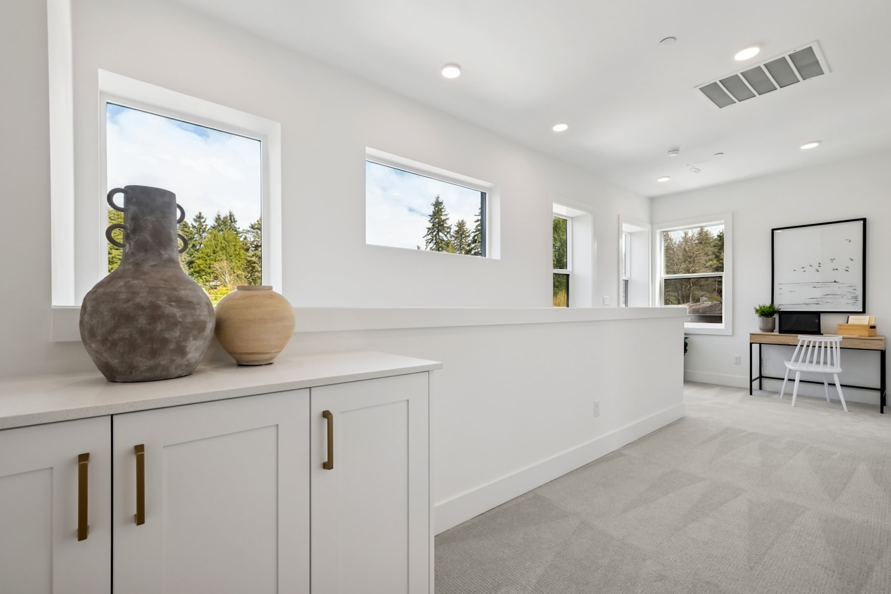 A modern, minimalist kitchen with white cabinets, wooden accents, and large windows overlooking a forested landscape.