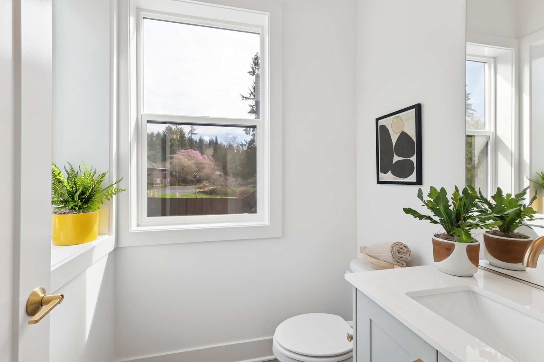 A bright and airy bathroom with a large window overlooking a scenic outdoor landscape, featuring potted plants and minimalist decor.
