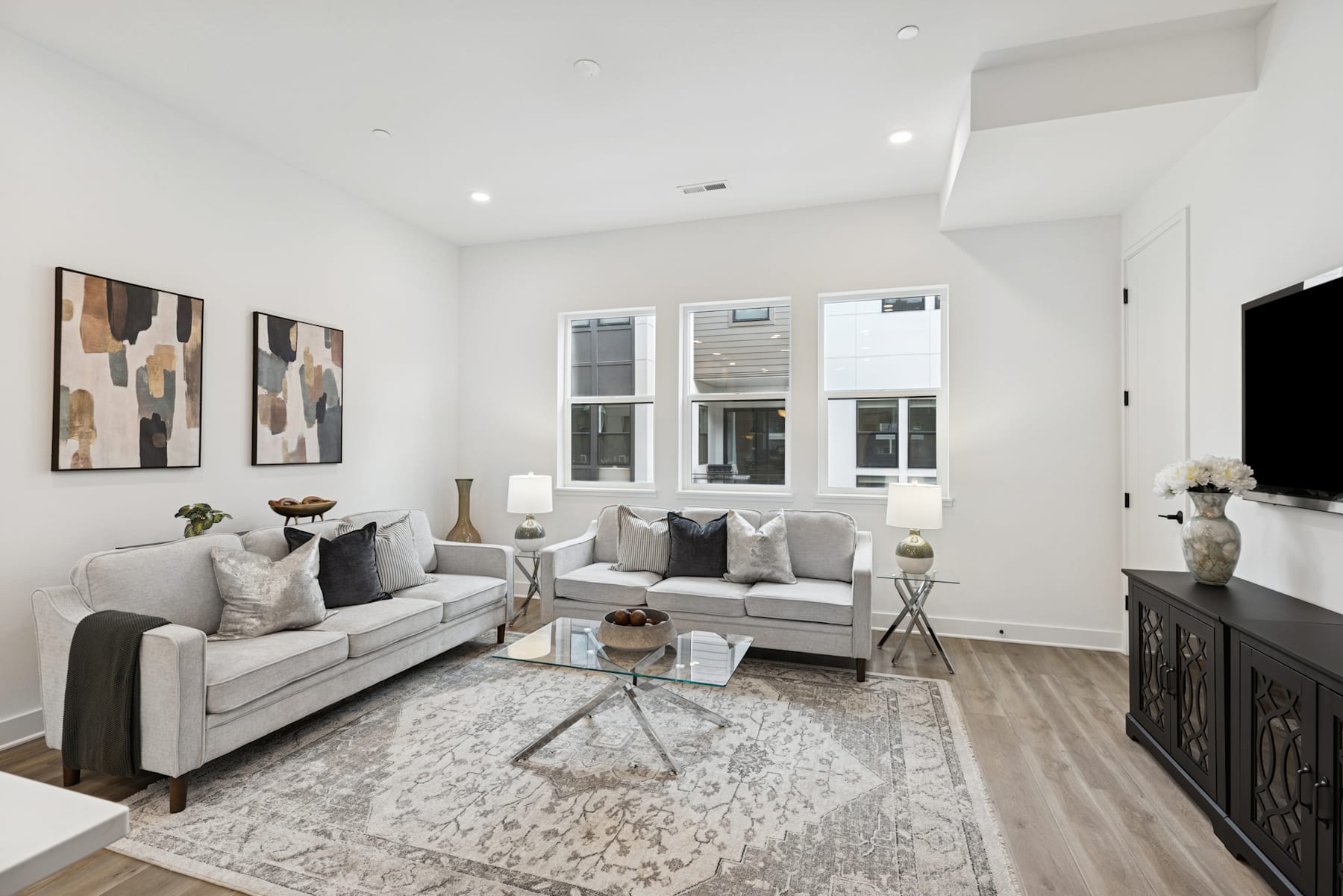 A modern and minimalist living room with a gray sofa, decorative pillows, and abstract artwork on the walls, set against a backdrop of large windows and hardwood flooring.