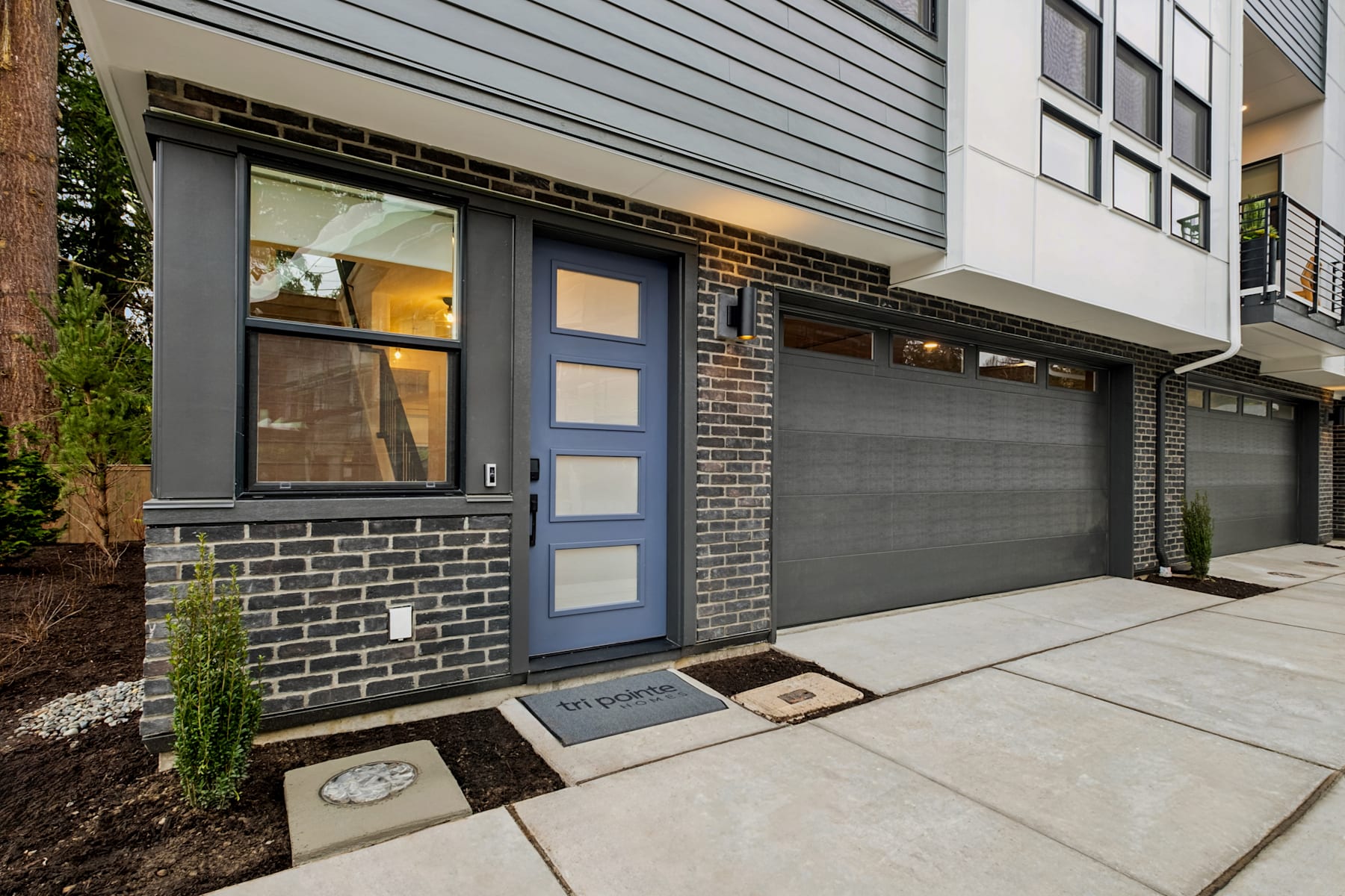 A modern, two-story residential building with a gray exterior, a garage door, and a blue front door surrounded by a brick facade, set against a backdrop of greenery.