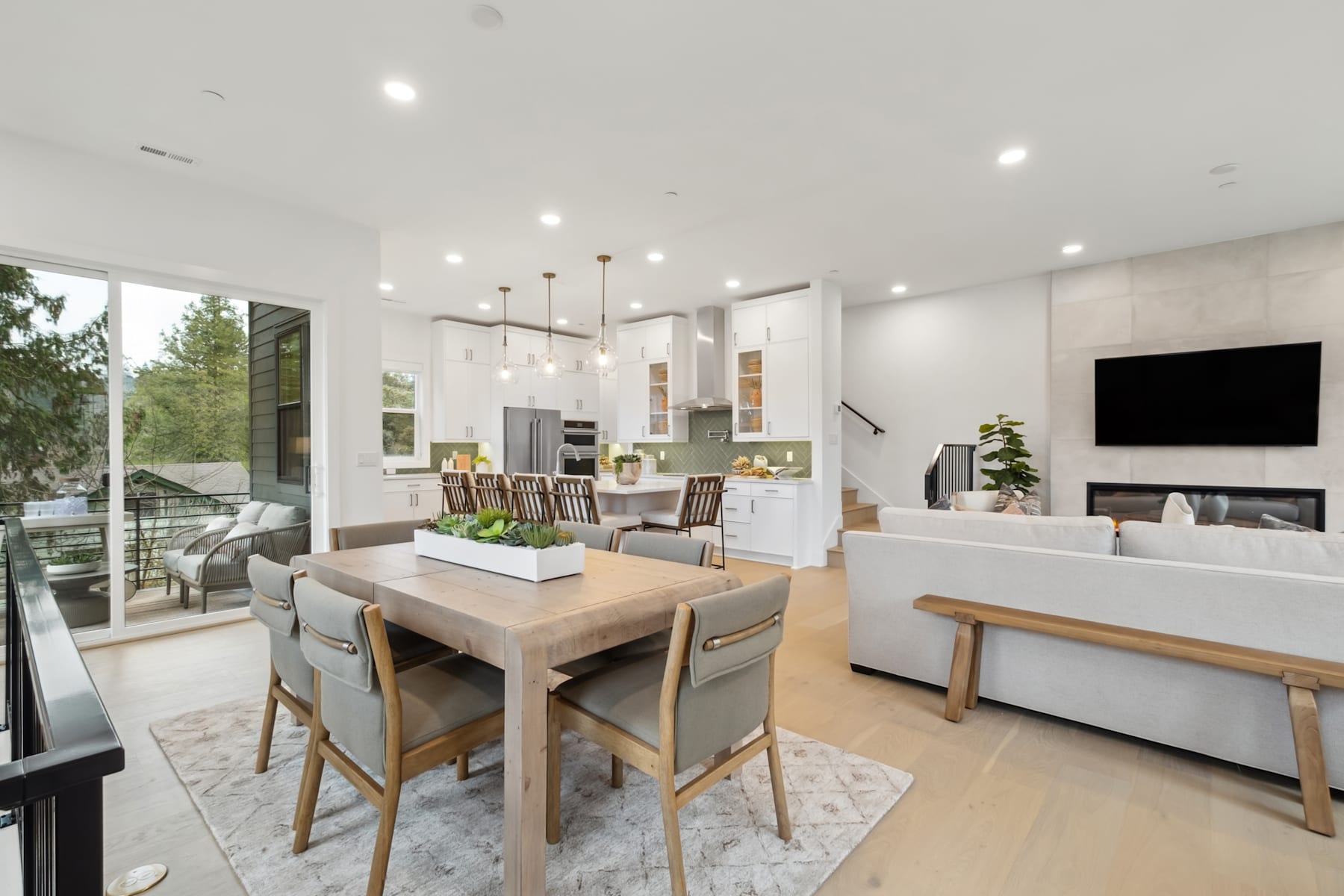 A modern and spacious open-concept living and dining area with a wooden dining table, comfortable seating, and large windows overlooking a natural outdoor setting.