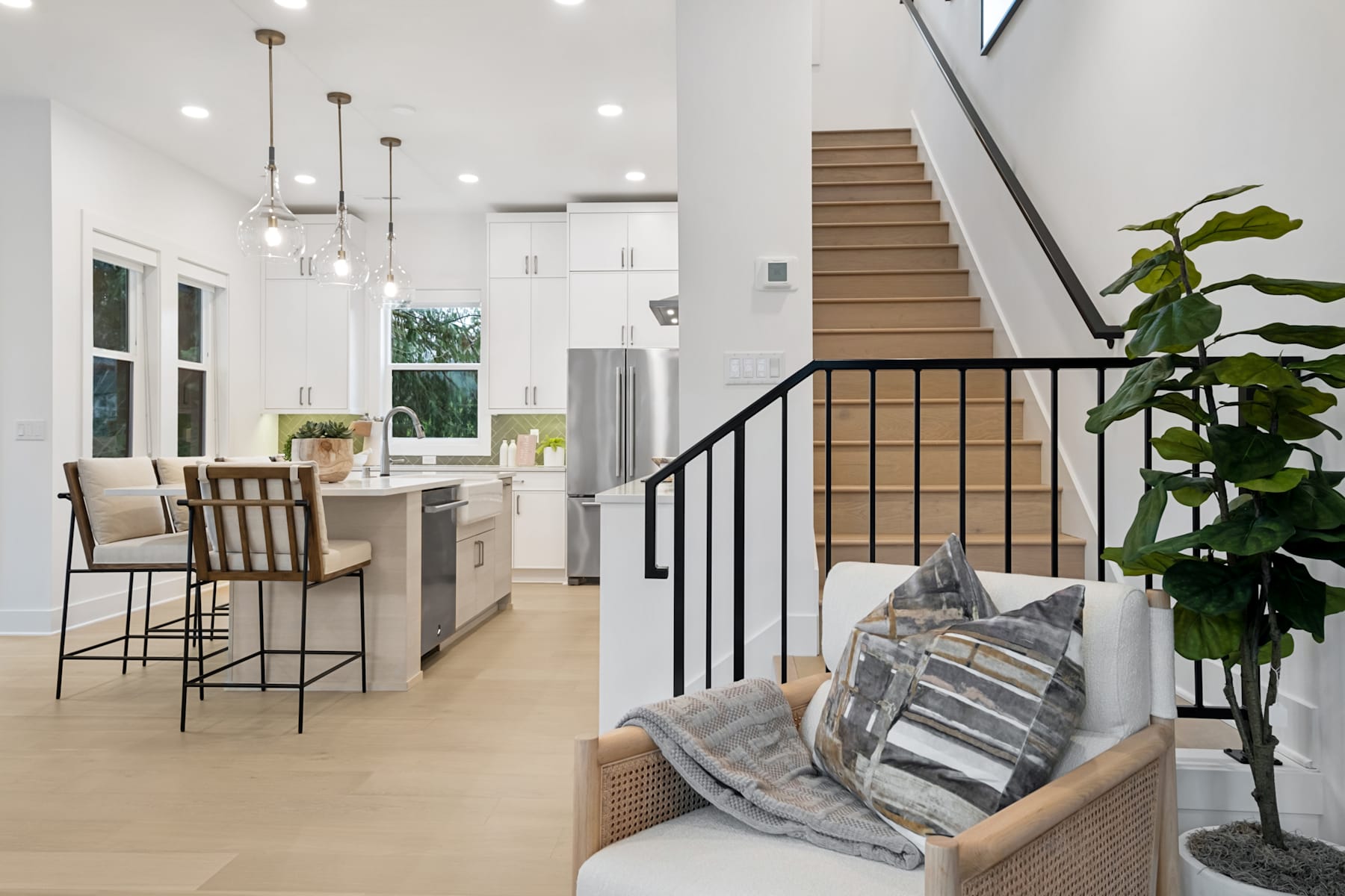 A modern and open-concept kitchen and living room with a staircase leading to an upper level, featuring a cozy seating area, potted plants, and a sleek, minimalist design.