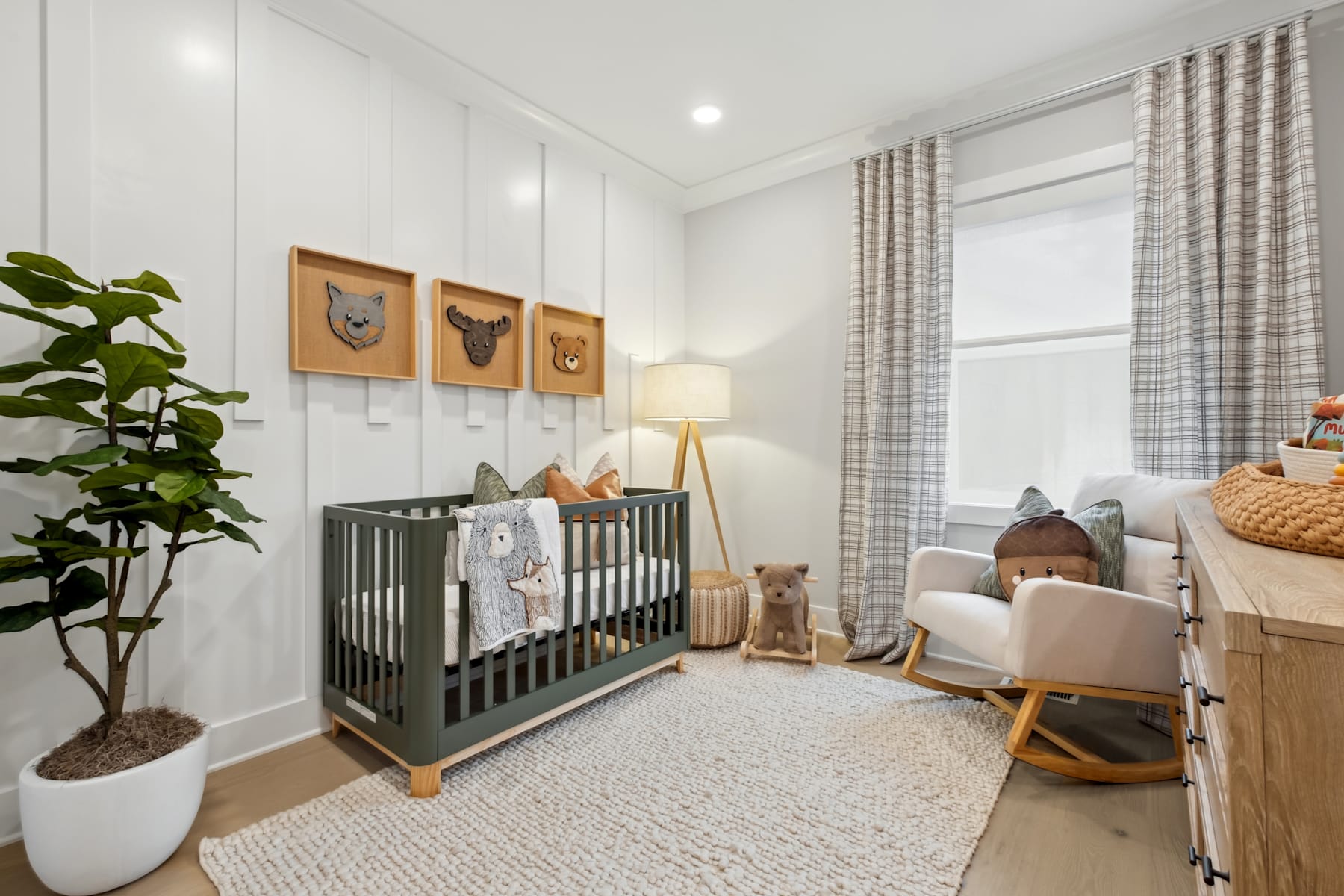 A cozy and well-decorated nursery with a crib, framed artwork, and a potted plant, set against a neutral-toned background with curtains and a comfortable seating area.