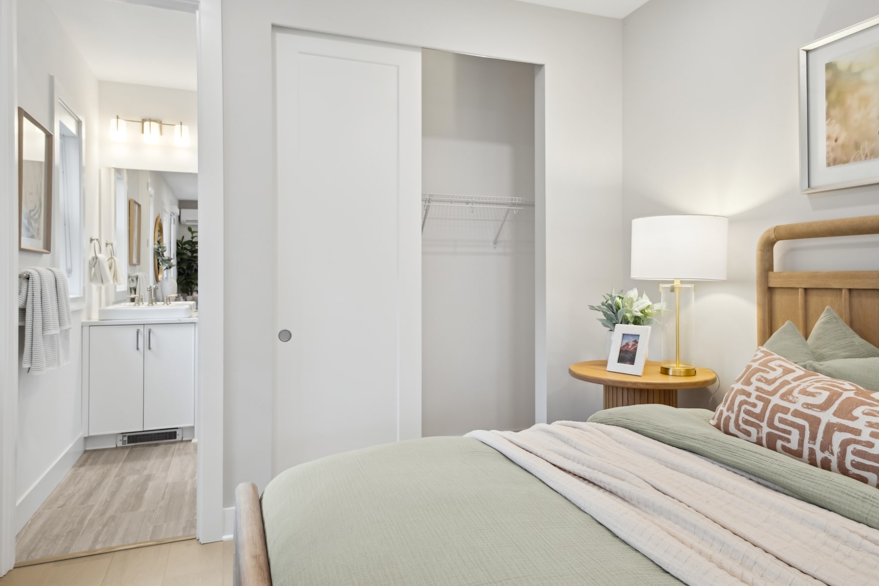 A cozy and well-lit bedroom with a wooden headboard, a side table with a lamp, and a framed artwork on the wall, leading to a hallway with a white door.