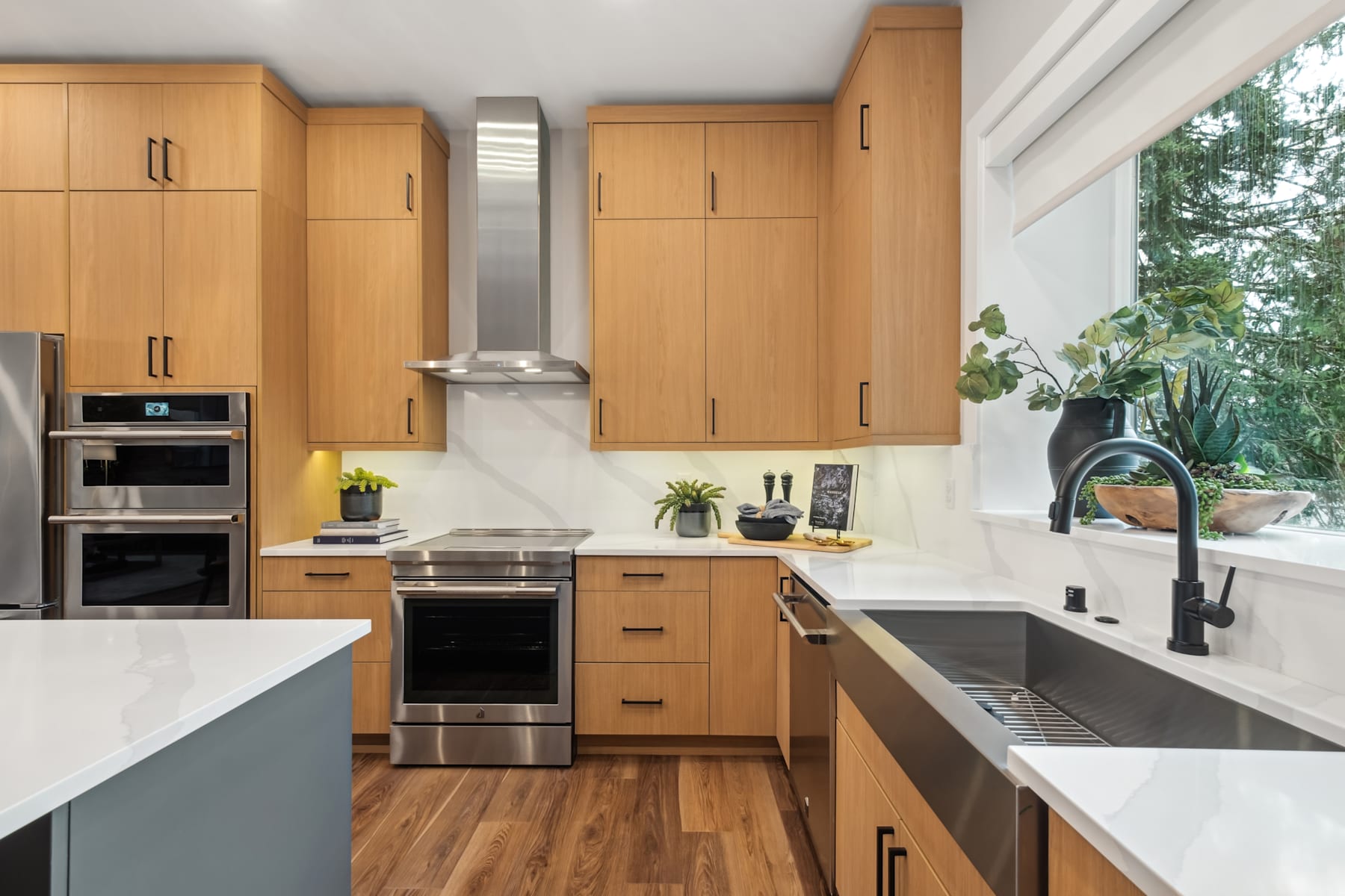 A modern and well-equipped kitchen with light wood cabinets, stainless steel appliances, and a large window overlooking a lush, forested outdoor area.