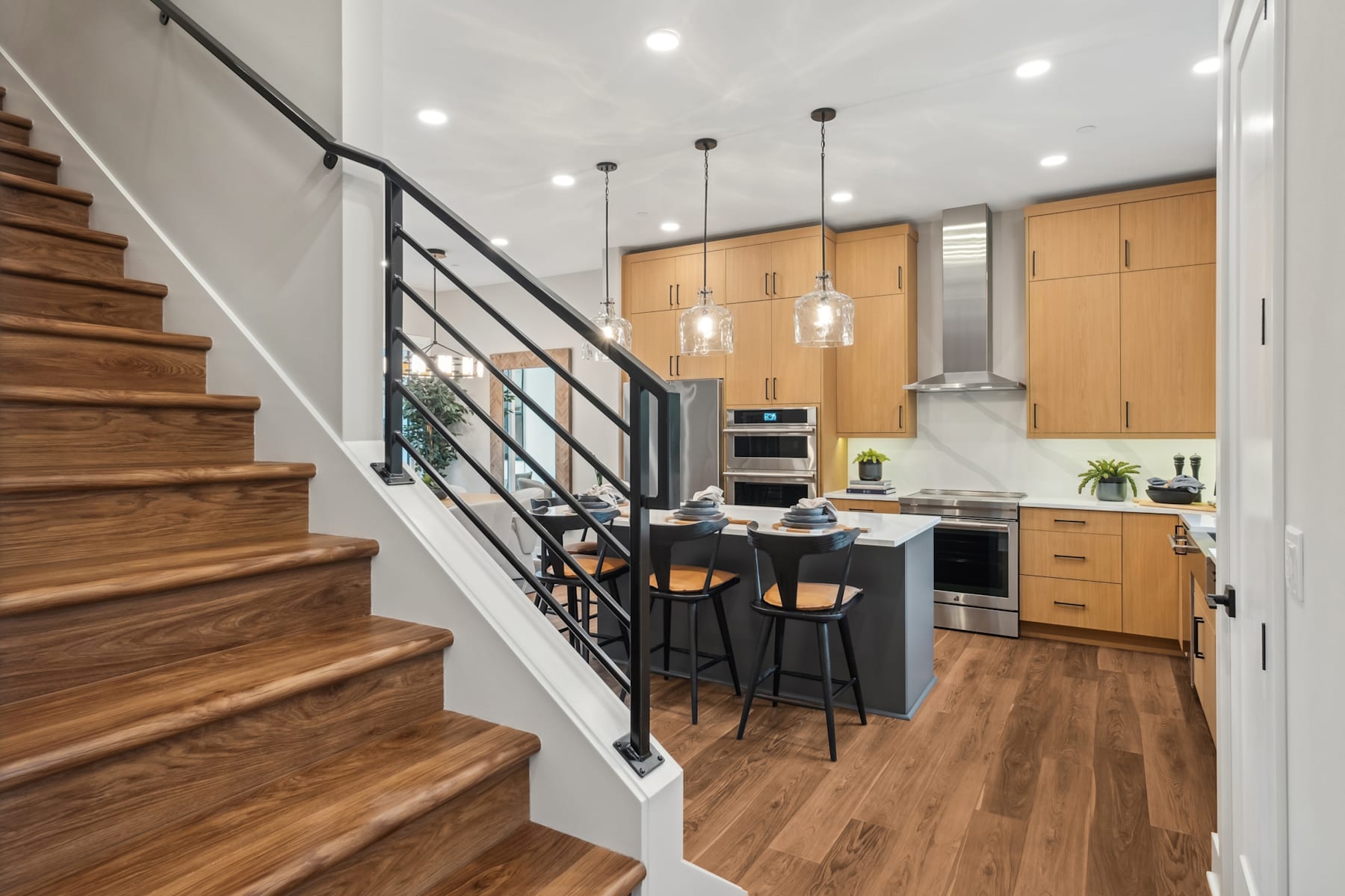 A modern kitchen with wooden stairs leading up to a second level, featuring sleek cabinetry, pendant lighting, and a central island with bar stools.