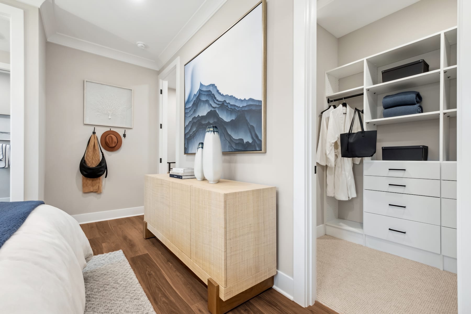 A modern and minimalist bedroom with a large painting of mountains on the wall, a wooden dresser, and a built-in closet system with shelves and drawers.