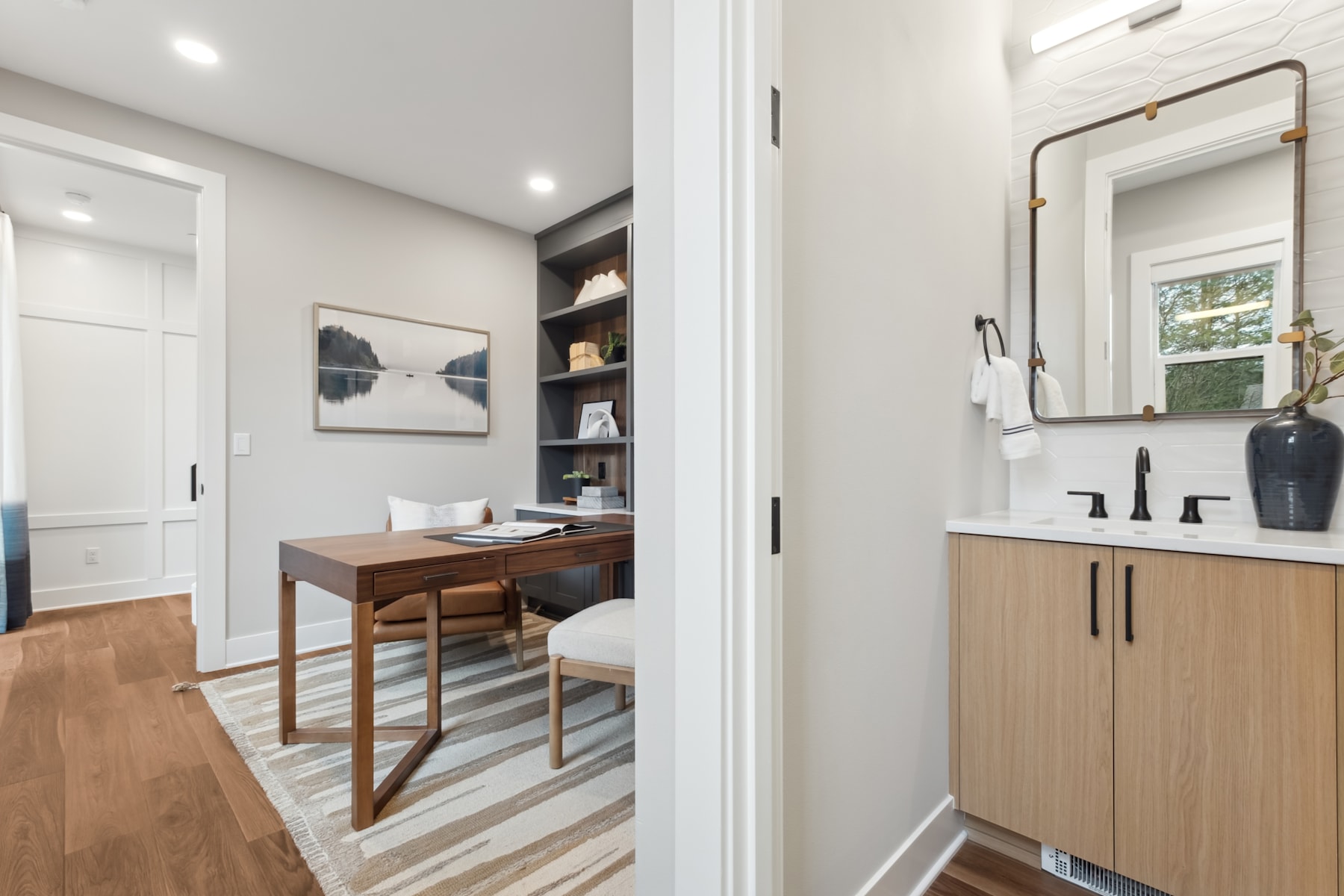 A modern and minimalist home office space with a wooden desk, shelves, and a bathroom vanity visible in the background.