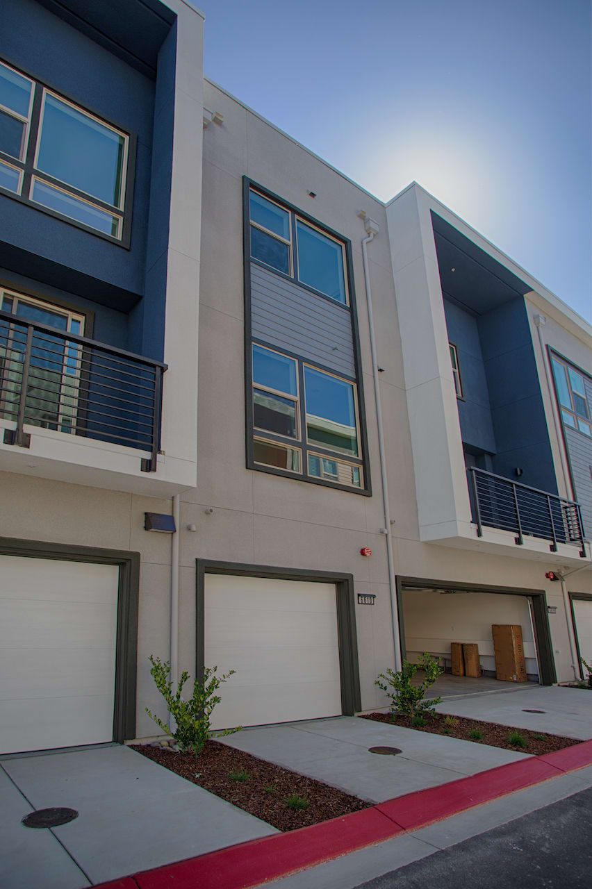 Modern multi-story townhouses with clean lines, large windows, and a mix of neutral and blue tones, set against a clear blue sky. The foreground features a paved driveway and landscaping with small plants.