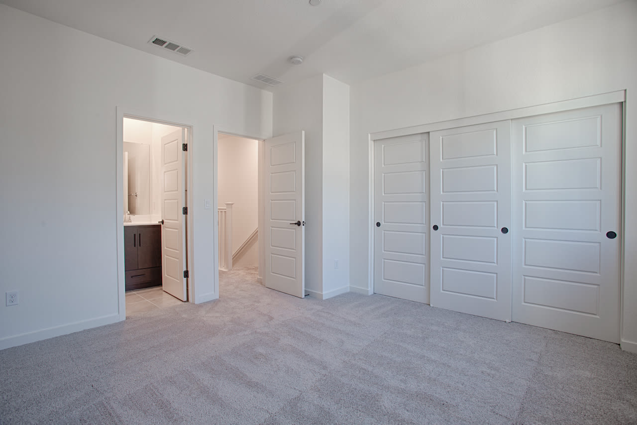 A spacious, minimalist bedroom with white walls, gray carpeting, and multiple closet doors in the foreground.