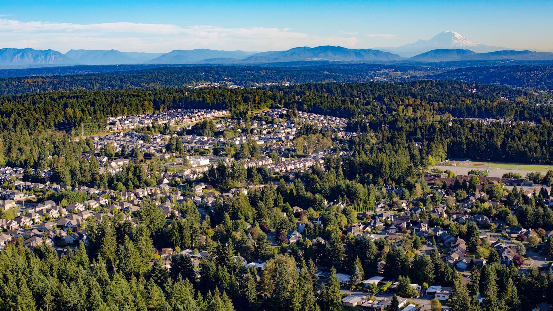 A lush, forested landscape with a small town nestled among the trees, surrounded by towering mountains in the distance under a clear blue sky.