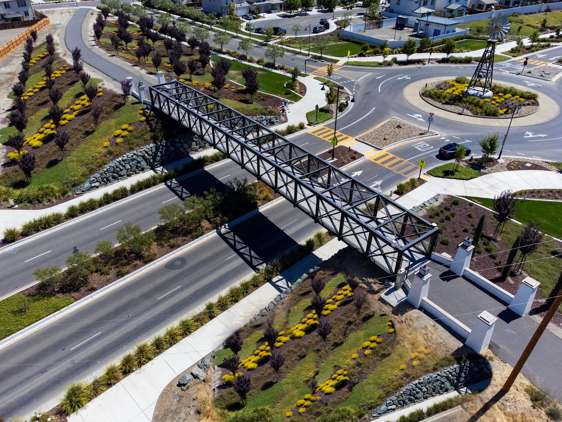 A large metal bridge spans across a road, surrounded by a landscaped area with trees and flowers in the foreground, and residential buildings in the background.