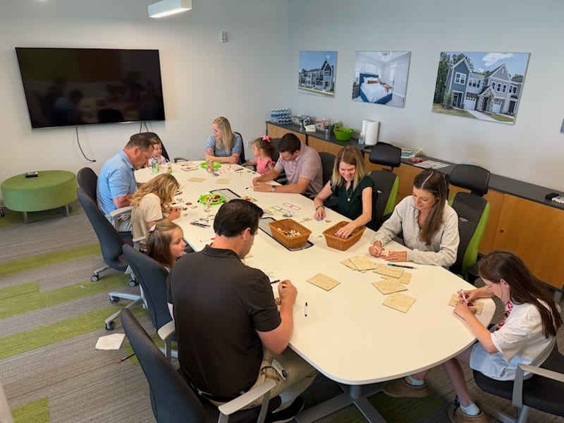 A group of people, including adults and children, are gathered around a large table, engaged in a collaborative activity or meal.