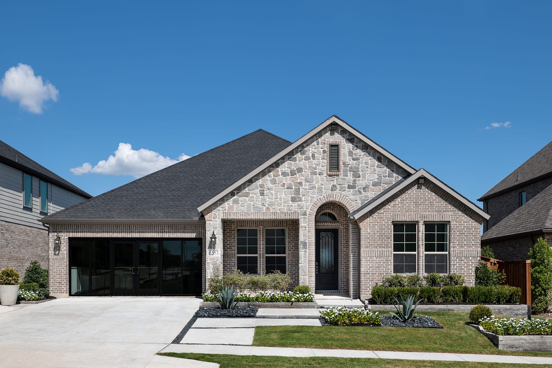 A well-designed, two-story brick house with a gabled roof, surrounded by a neatly landscaped yard featuring a paved driveway and lush greenery against a clear blue sky with scattered clouds.