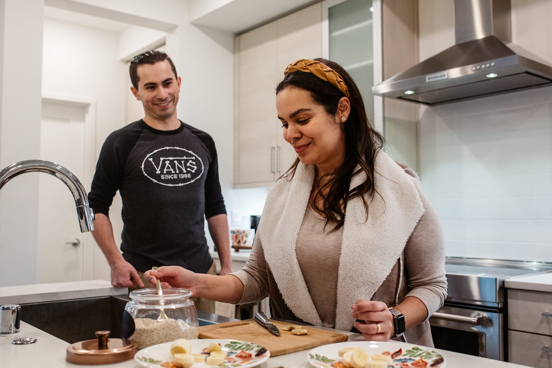 A young couple is preparing food together in a modern kitchen, with the woman wearing a headband and the man wearing a Vans t-shirt.