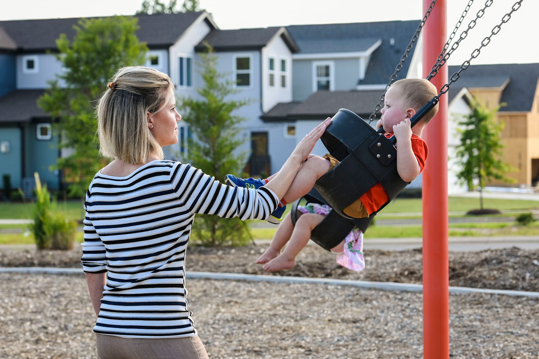 A woman in a striped shirt is pushing a young child on a swing in a playground surrounded by residential buildings.