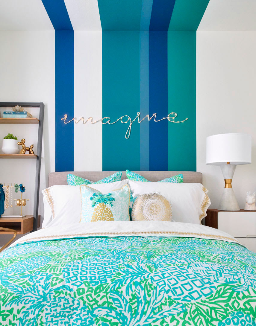 A vibrant and colorful bedroom with a patterned bedspread, shelving unit, and a wall featuring bold blue and green stripes with the word "imagine" written in cursive.