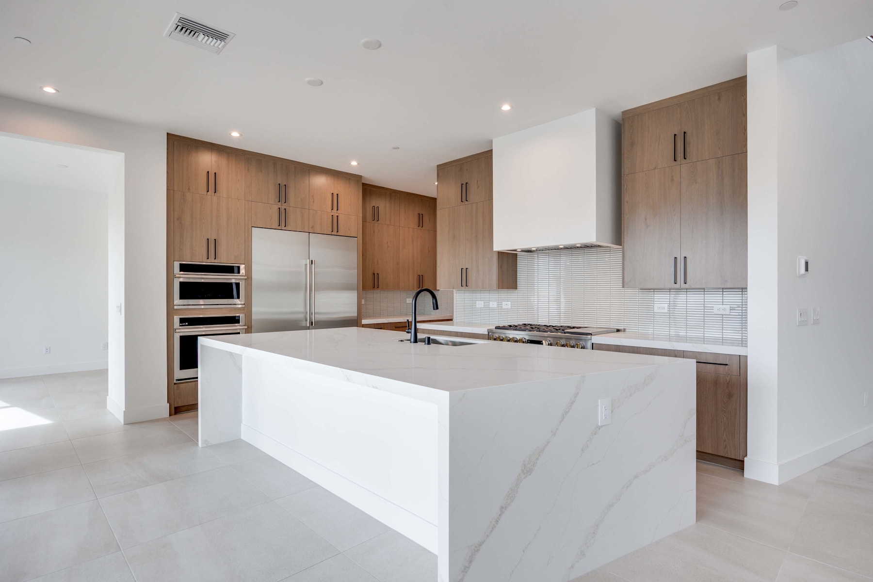 A modern, open-concept kitchen with a large white island, wooden cabinetry, and stainless steel appliances, set against a bright and airy background.