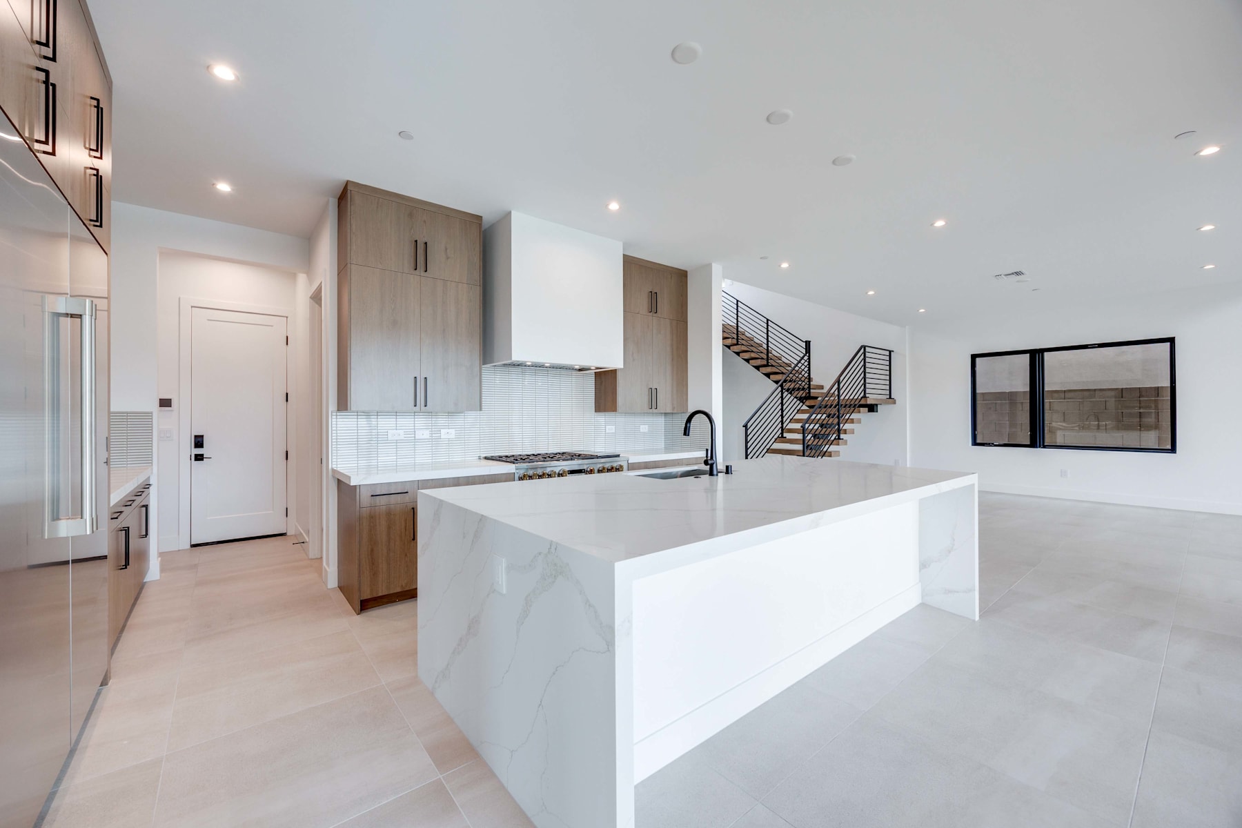 A modern, open-concept kitchen with a white island, wooden cabinets, and a staircase leading to an upper level in the background.