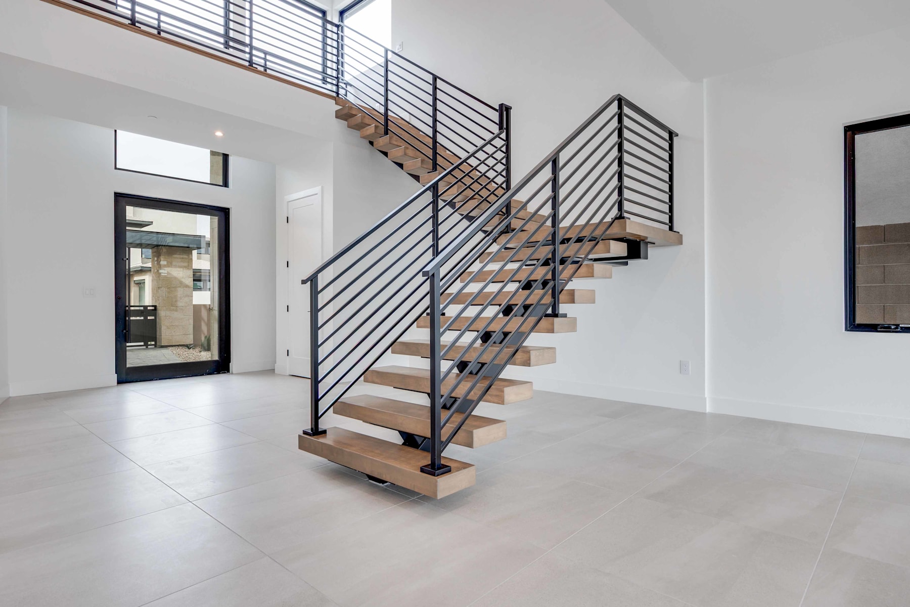 A modern, open-concept entryway with a wooden and metal staircase leading to an upper level, surrounded by white walls and a tiled floor.