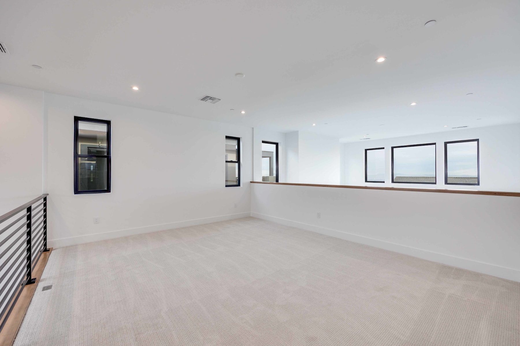 A spacious, minimalist room with white walls, hardwood floors, and large windows providing ample natural light. The room features a railing along the upper level, creating an open and airy atmosphere.