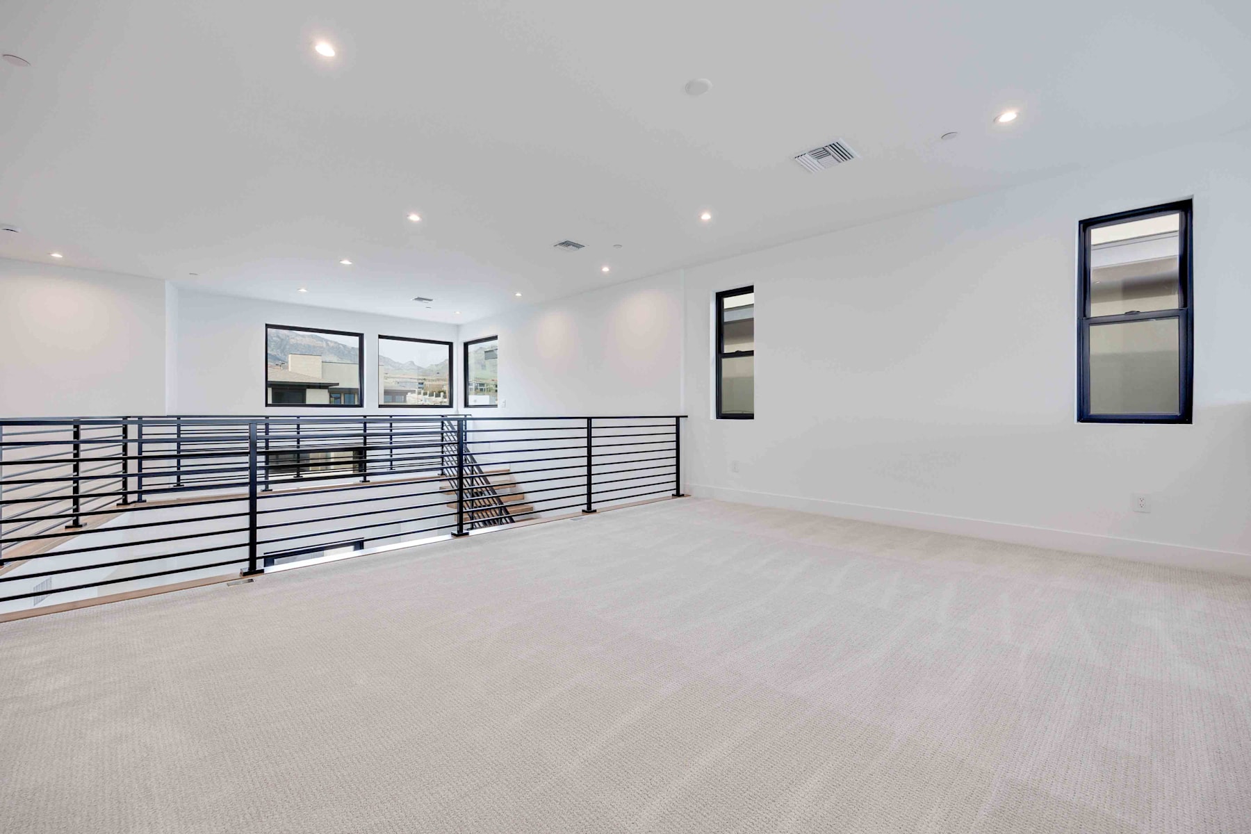 A spacious, open-concept room with a loft-style design, featuring a white floor, black metal railings, and large windows allowing natural light to flood the space.