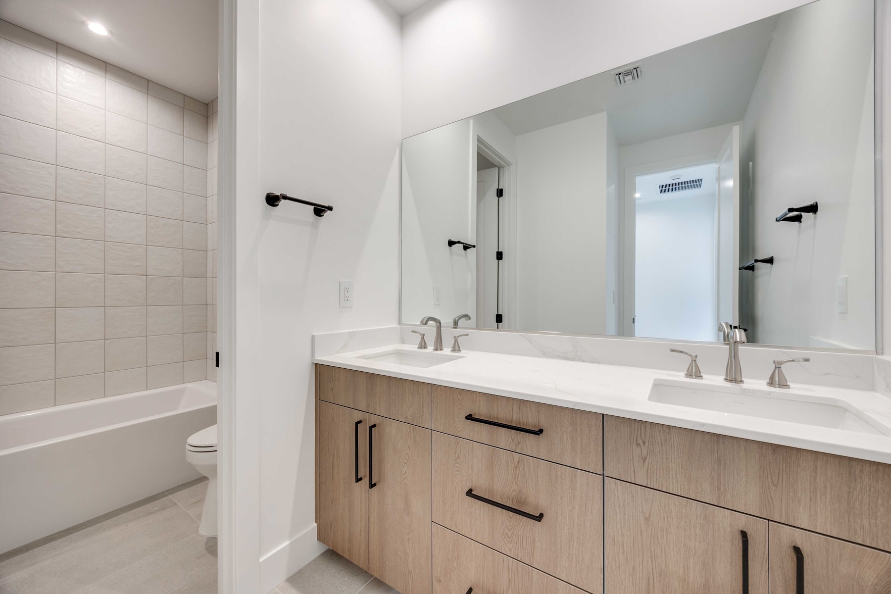 A modern bathroom with a double vanity, white countertop, and wood-toned cabinets, along with a large mirror and recessed lighting.
