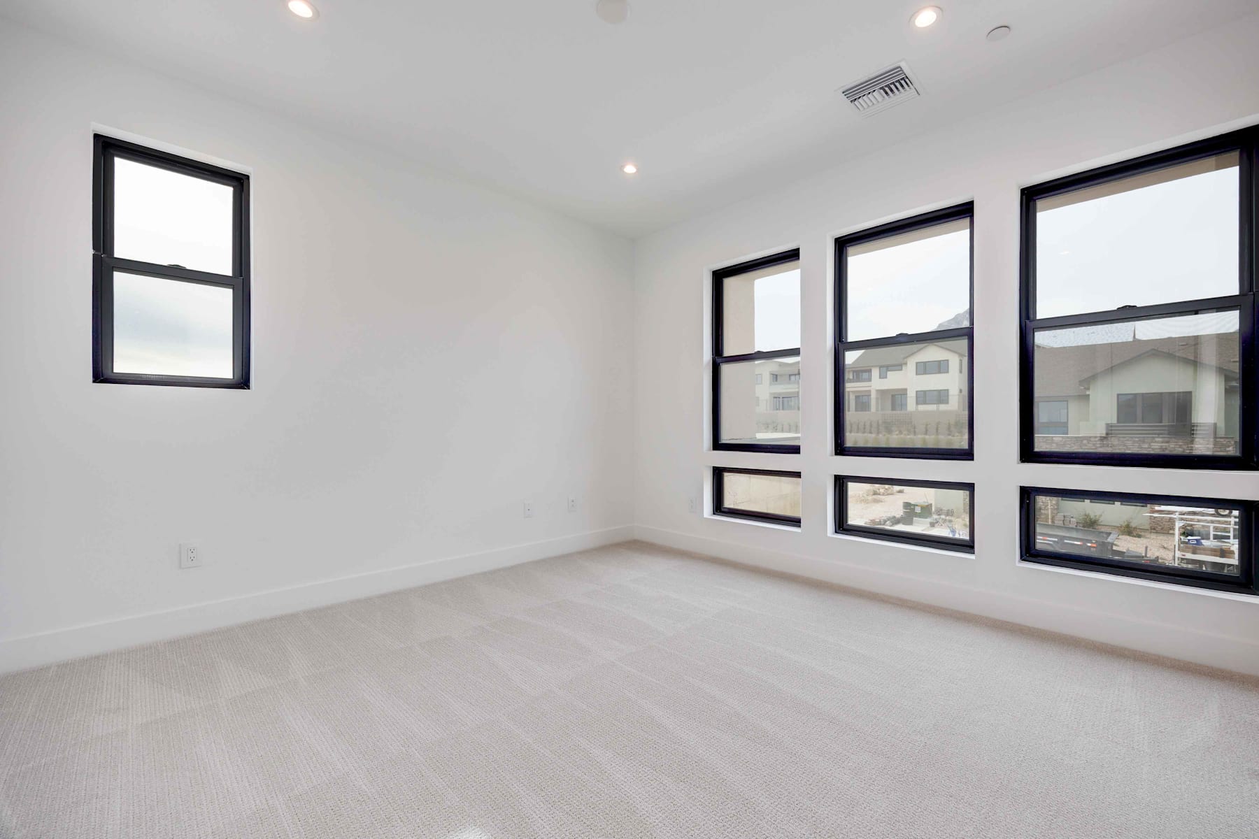A spacious, minimalist room with large windows providing ample natural light, a white floor, and black-framed windows overlooking the surrounding buildings.