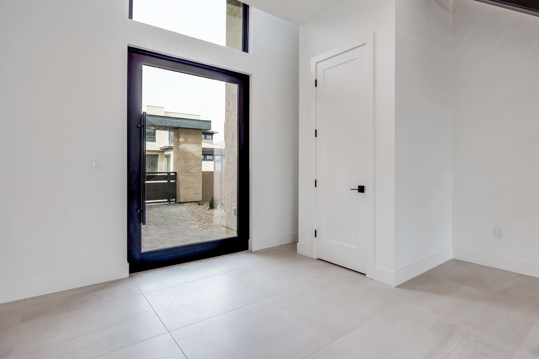 A spacious, minimalist interior with a large window, a white door, and a tiled floor, creating a bright and airy atmosphere.