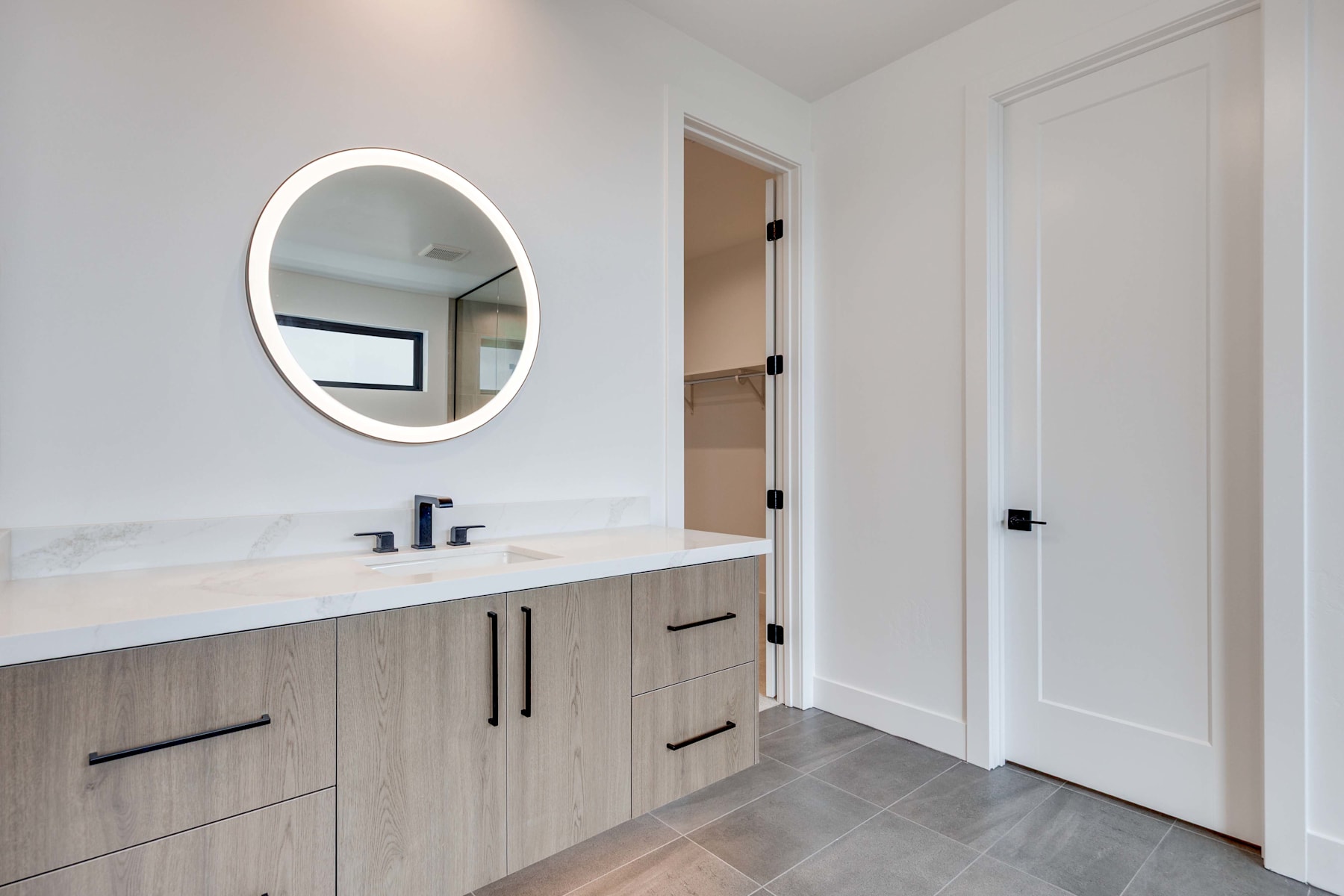 A modern bathroom with a vanity, oval mirror, and a doorway leading to another room.