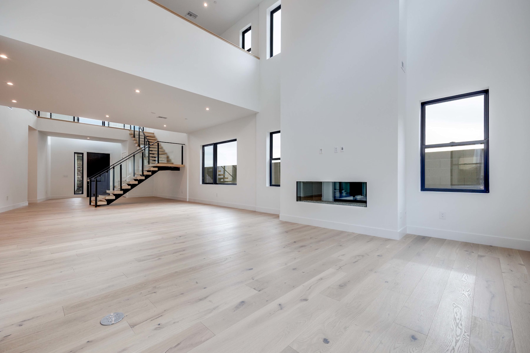 A spacious, modern interior with a high ceiling, hardwood floors, and a staircase leading to an upper level. The room appears to be a living or dining area, with a television mounted on the wall.