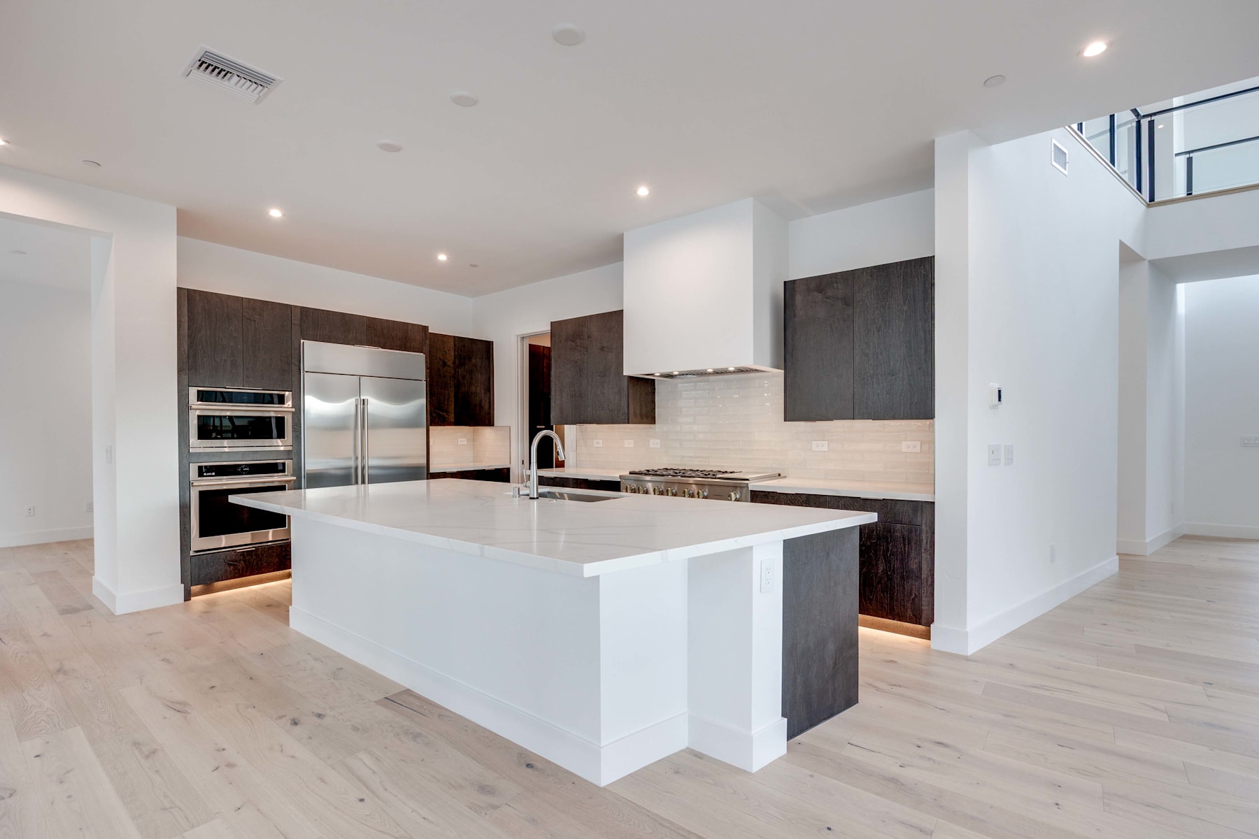 A modern, open-concept kitchen with a large white island, stainless steel appliances, and dark wood cabinetry, set against a bright and airy background.