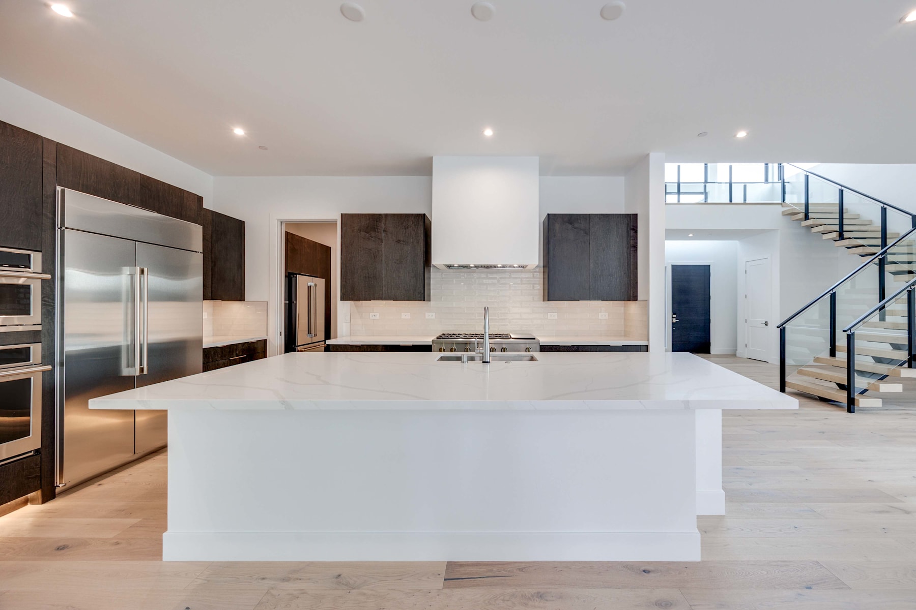 A modern, open-concept kitchen with a large white island, stainless steel appliances, and dark wood cabinetry, set against a bright and airy backdrop with a staircase visible in the background.