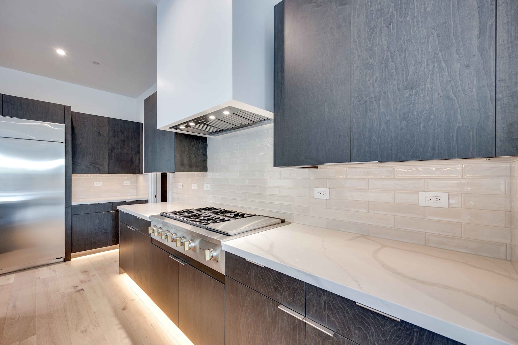 A modern kitchen with dark cabinets, a gas stove, and a sleek range hood, set against a backdrop of light-colored tile backsplash and countertops.