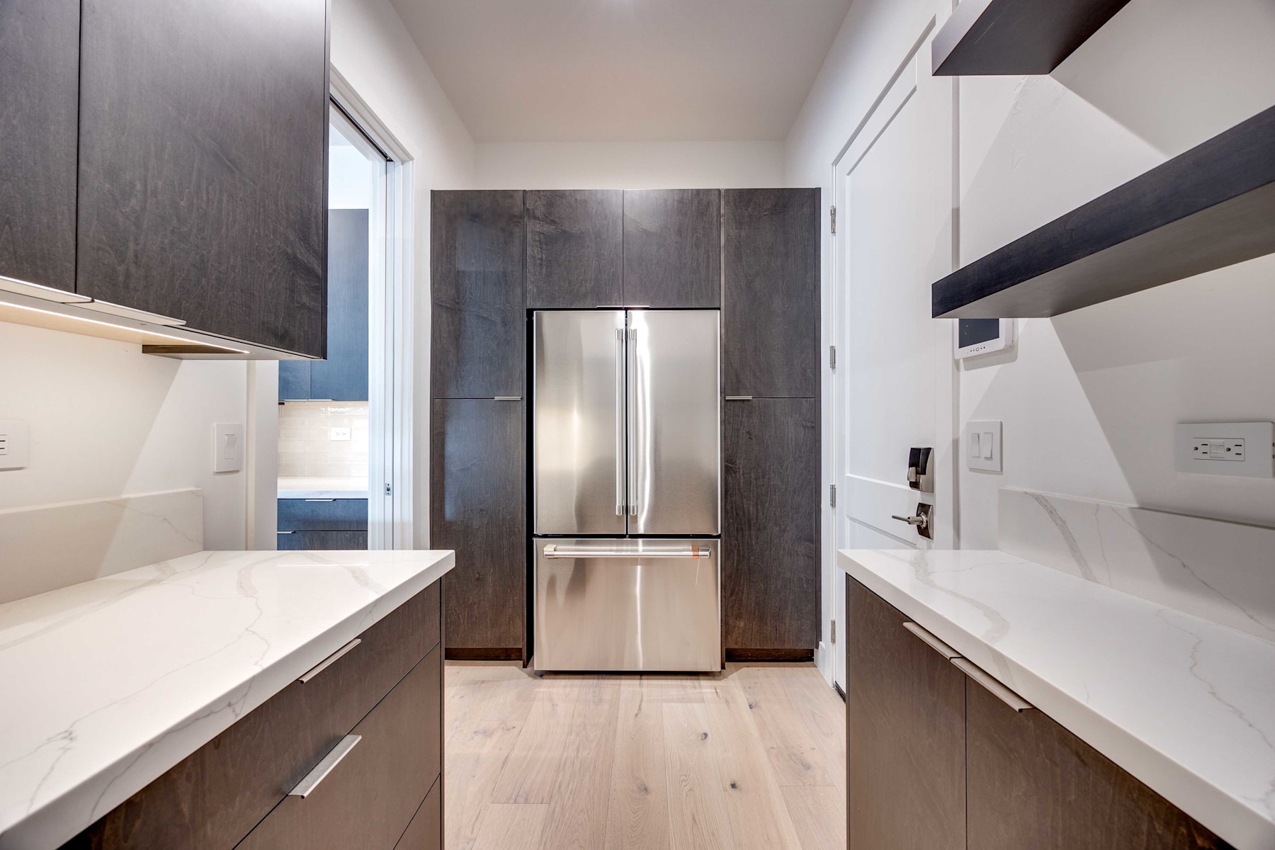 A modern, minimalist kitchen with sleek white countertops, dark cabinets, and a stainless steel refrigerator in the background.