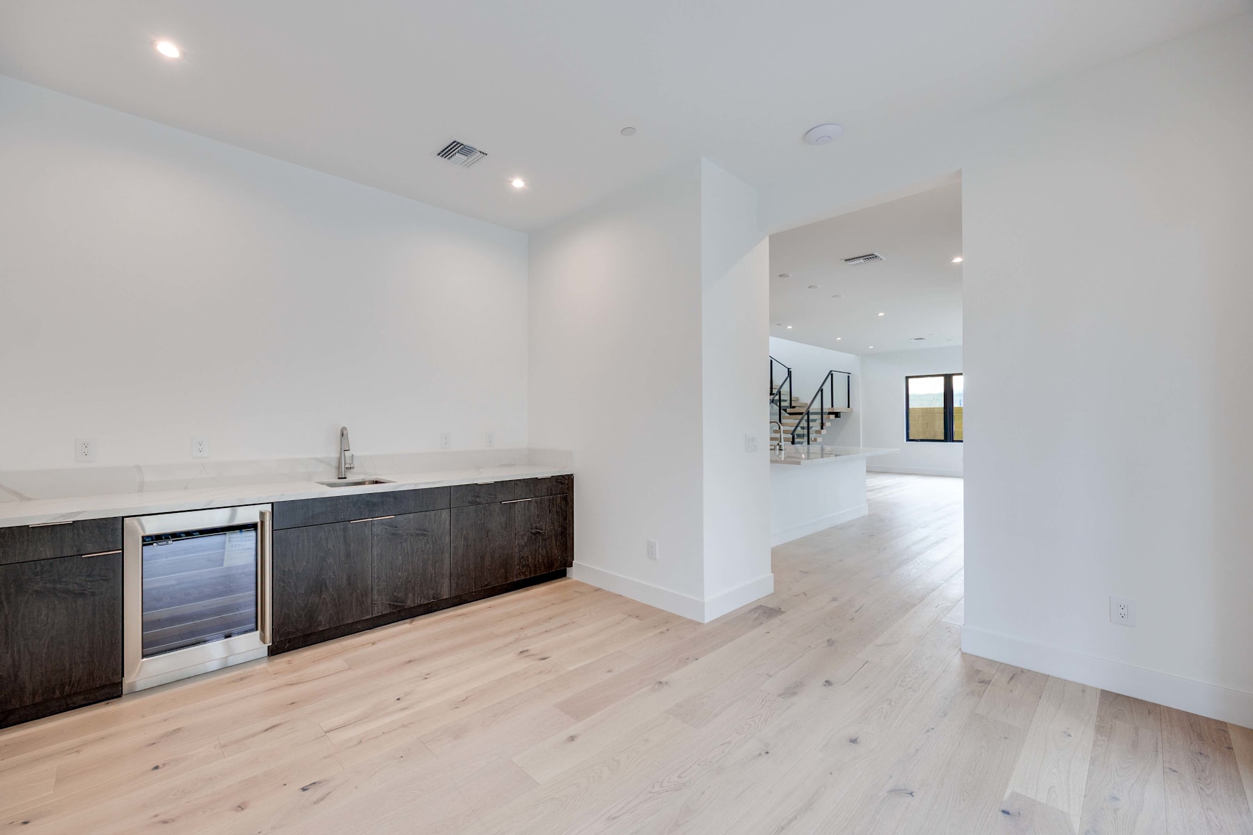 A modern, open-concept kitchen with dark wood cabinets and light hardwood floors, leading into a bright, airy living space.