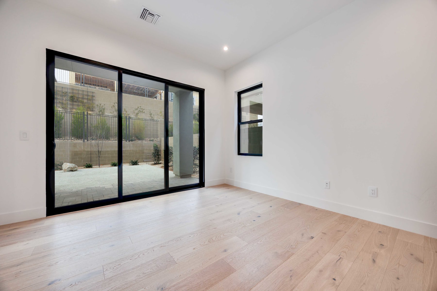 A spacious, minimalist room with large sliding glass doors leading to an outdoor area, featuring hardwood floors and a simple, clean aesthetic.