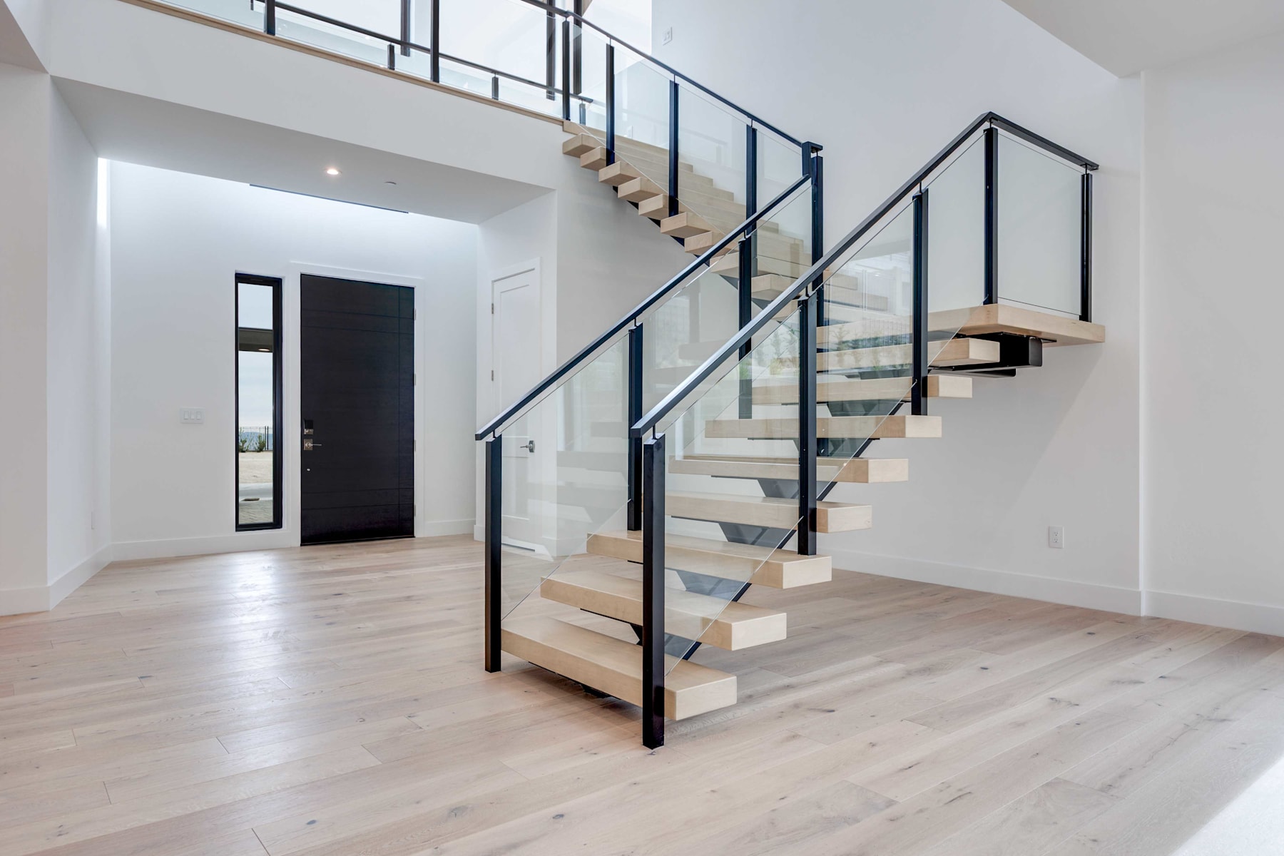 A modern, minimalist staircase with wooden steps and a sleek, black metal railing stands in a bright, open-plan interior with white walls and hardwood flooring.